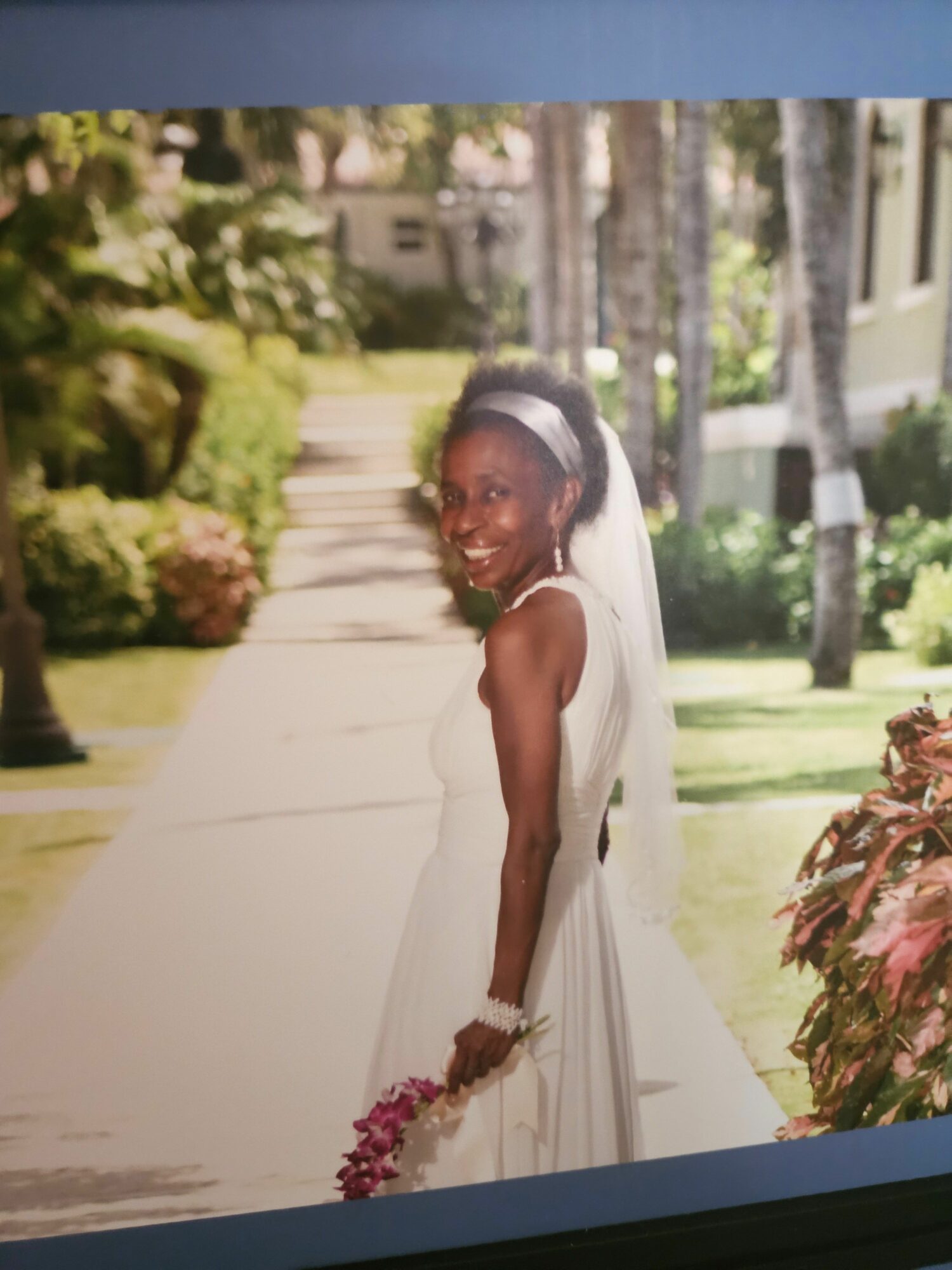 Woman in white dress holding flowers outdoors, with trees and buildings in background.