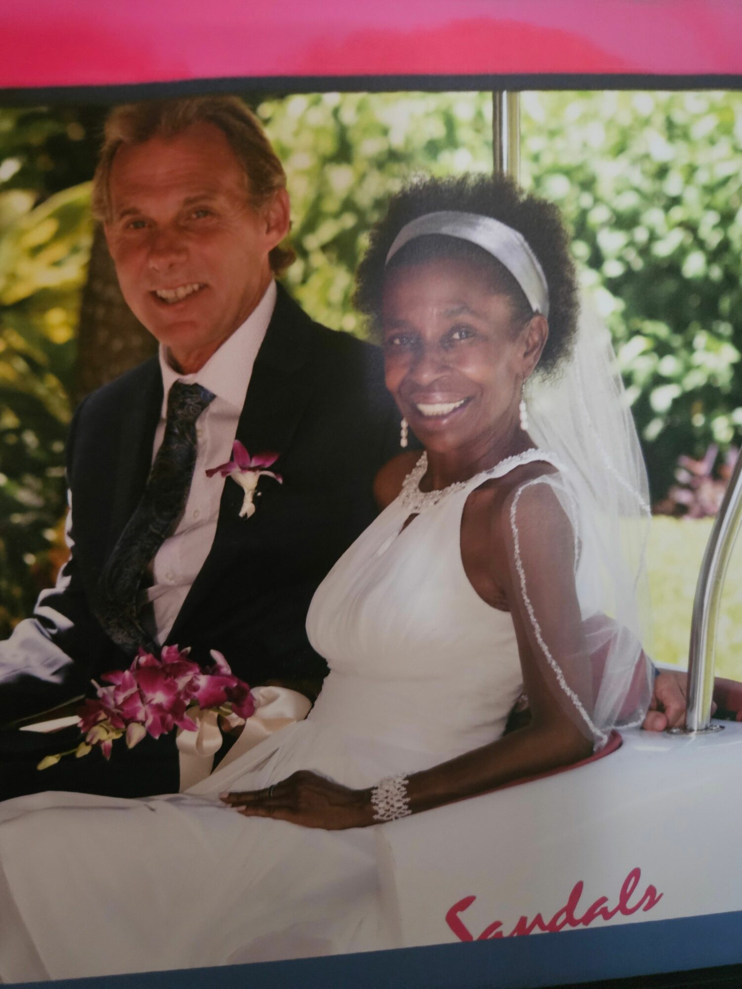 Bride and groom smiling at wedding, bride in white dress, groom in suit, outdoors with greenery background.