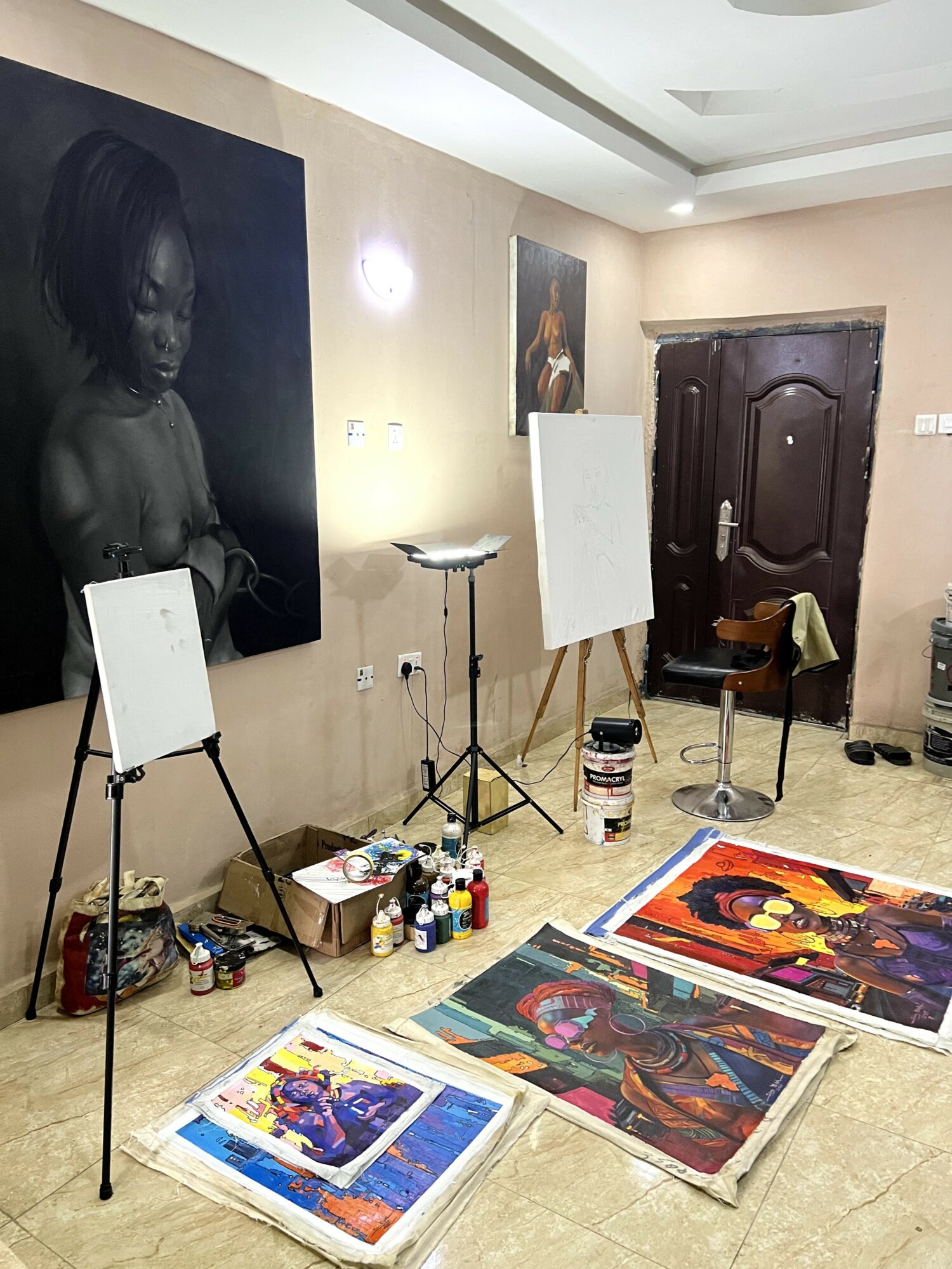 Art studio with paintings, easels, and supplies, including colorful paintings on the floor and a dark wooden door.