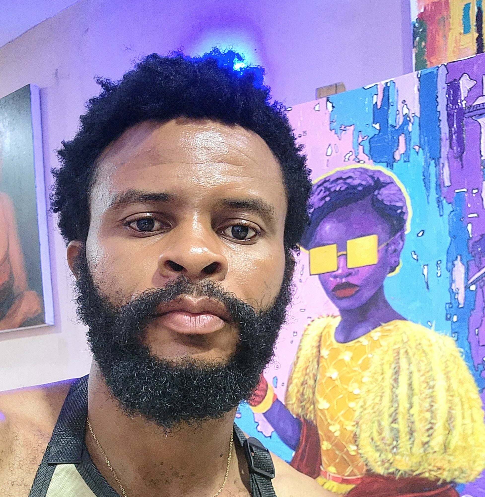 Man with curly hair and beard taking a selfie in front of colorful artwork featuring a person with sunglasses and yellow dress.