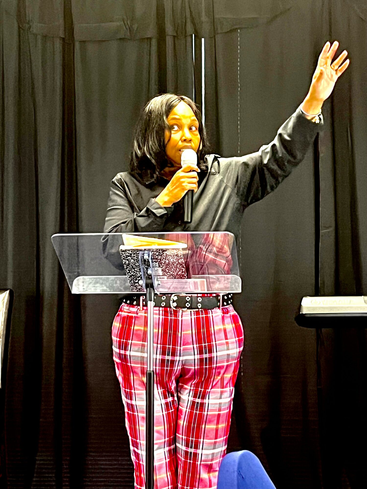 Woman speaking into microphone with her right arm raised, standing behind a clear podium, black curtain background.