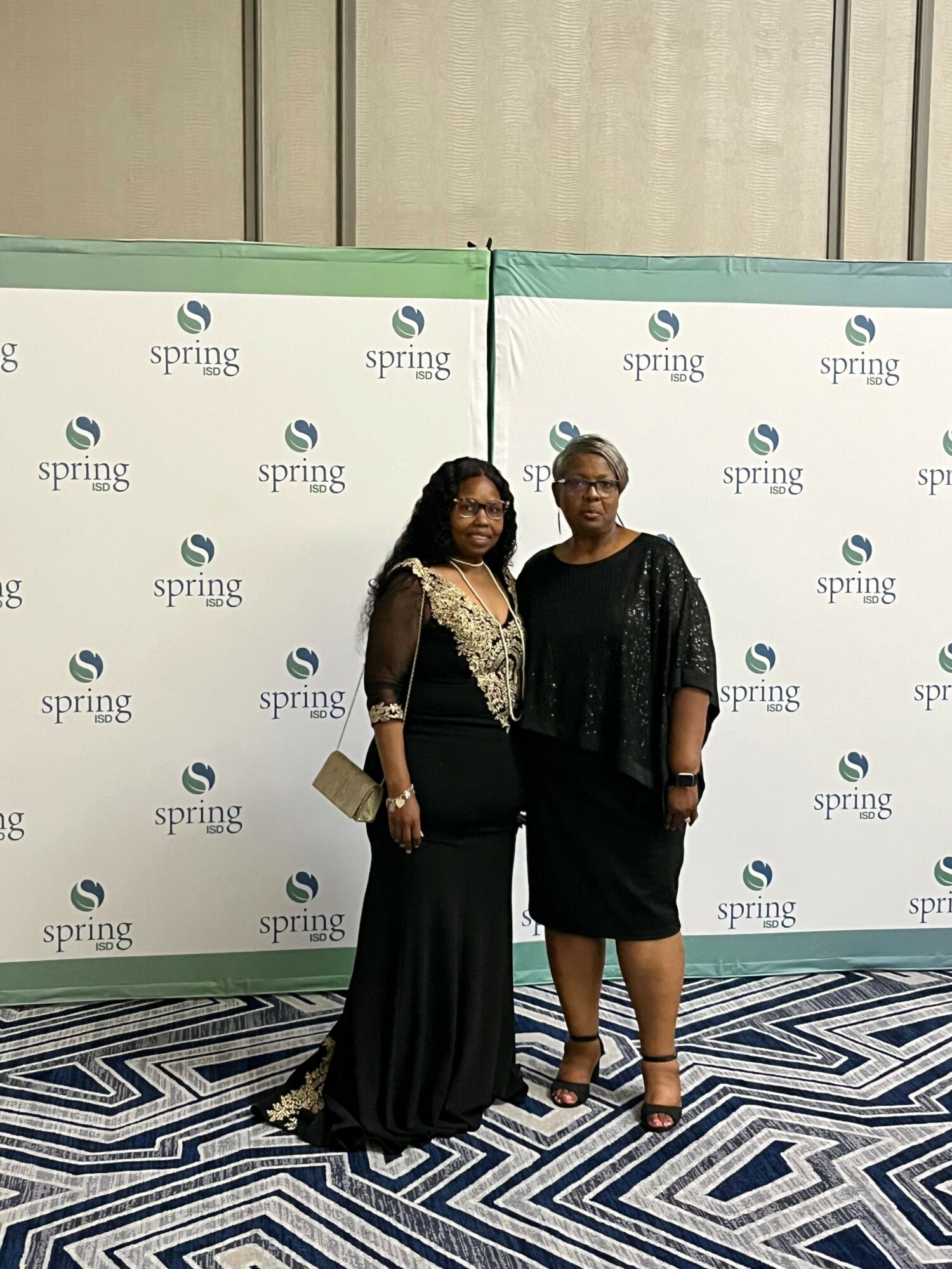 Two women standing side by side in front of a backdrop with a repeating logo pattern, on a patterned carpet.