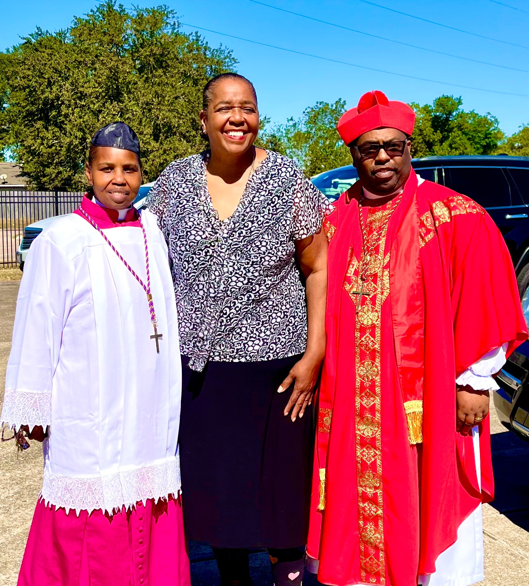 Three people standing outdoors, smiling, with trees and a car in the background. One person wears a white and pink religious robe, another in casual clothing, and the third in red ceremonial attire.