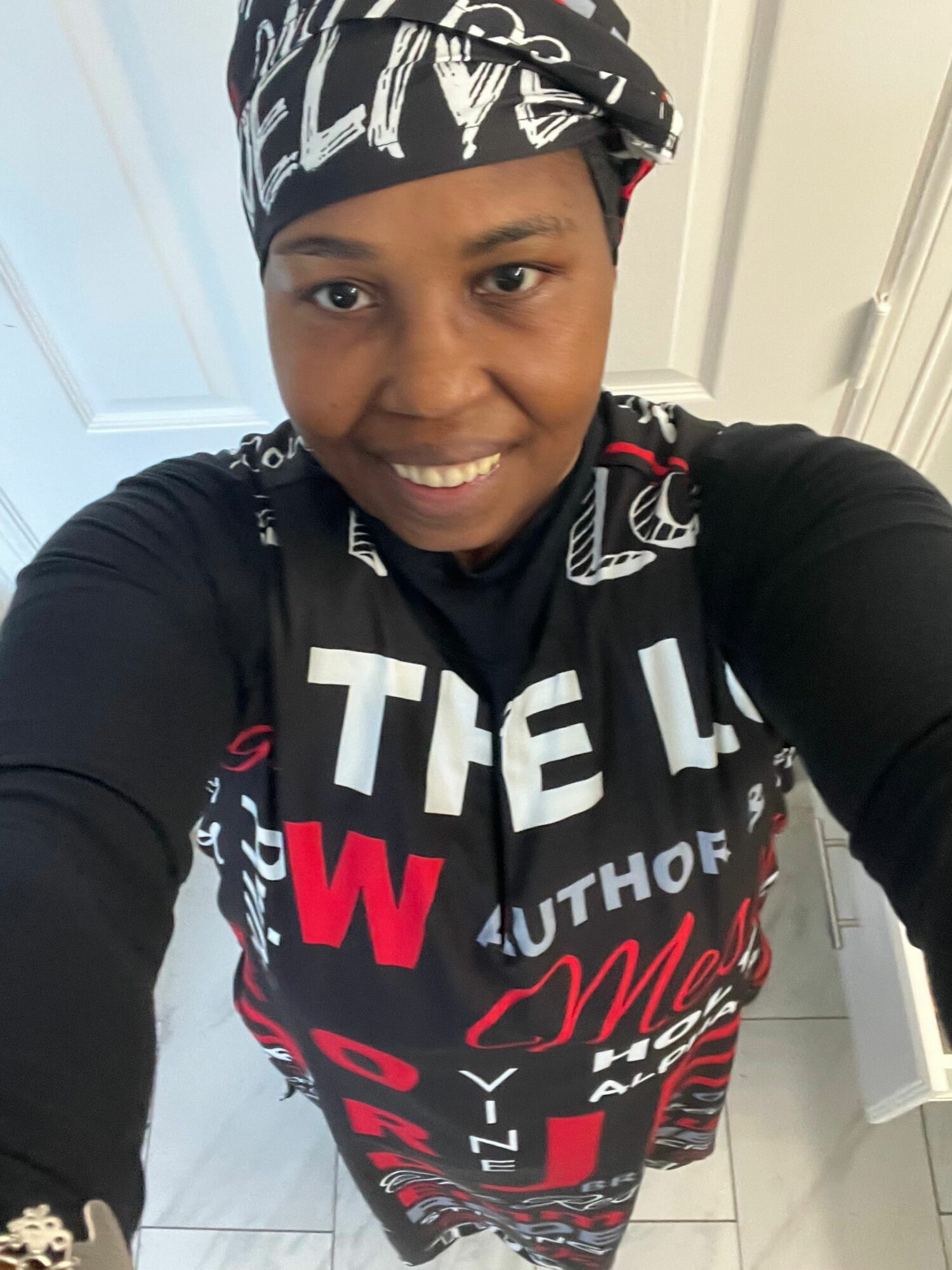 Smiling person wearing a black and white headscarf and a black shirt with red and white text, taking a selfie indoors.