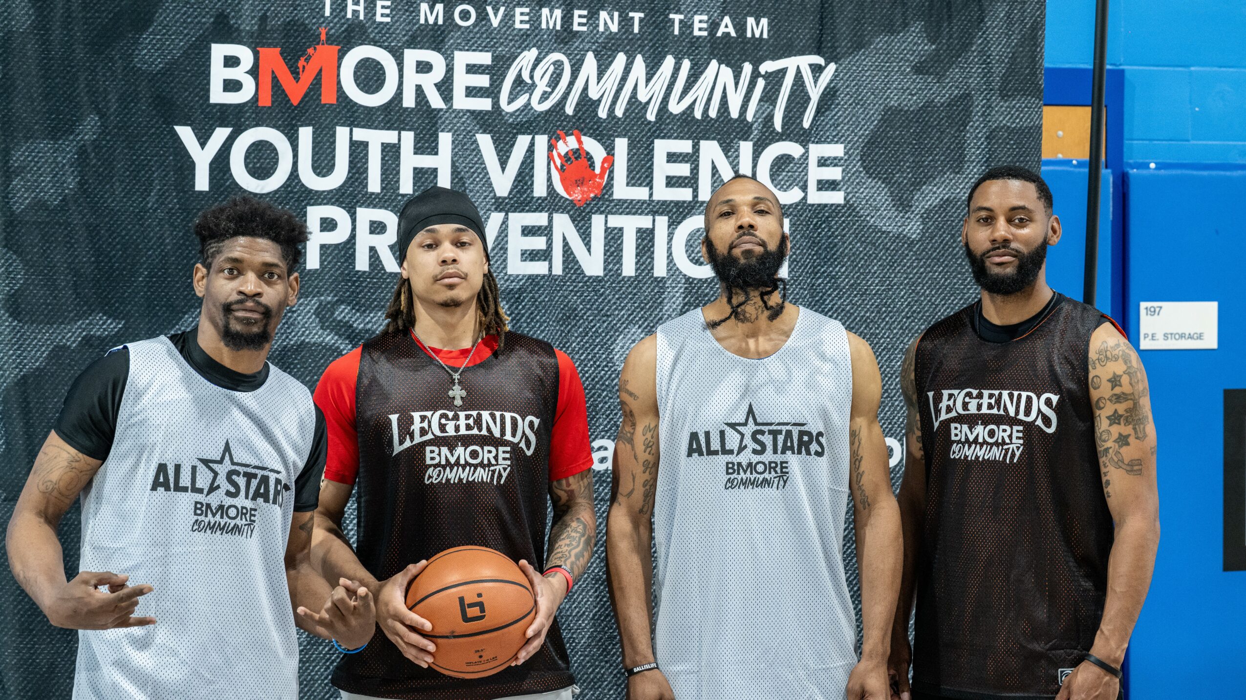 Four young men stand in front of a wall with text about youth violence prevention, some wearing basketball jerseys, one holding a basketball.