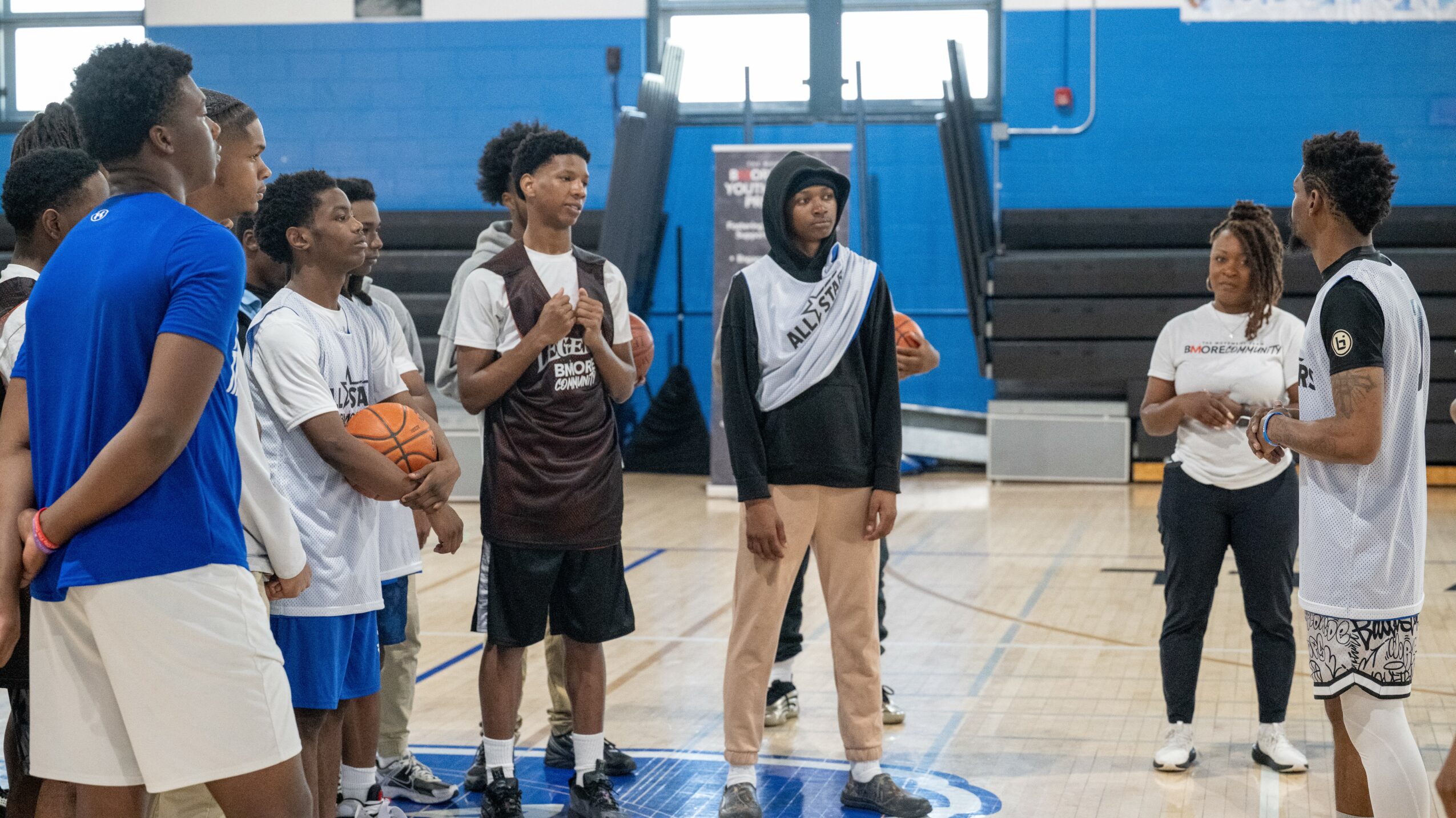 Group of people in a gymnasium, some holding basketballs, listening to a person speaking, with a woman holding a clipboard.
