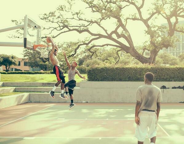 Two children playing basketball outdoors with a man watching, trees and buildings in the background, sunny day.