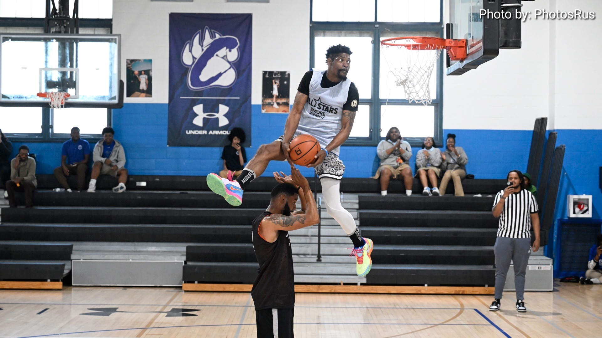 Basketball player mid-air dunking over another player in gymnasium with spectators and referee present.