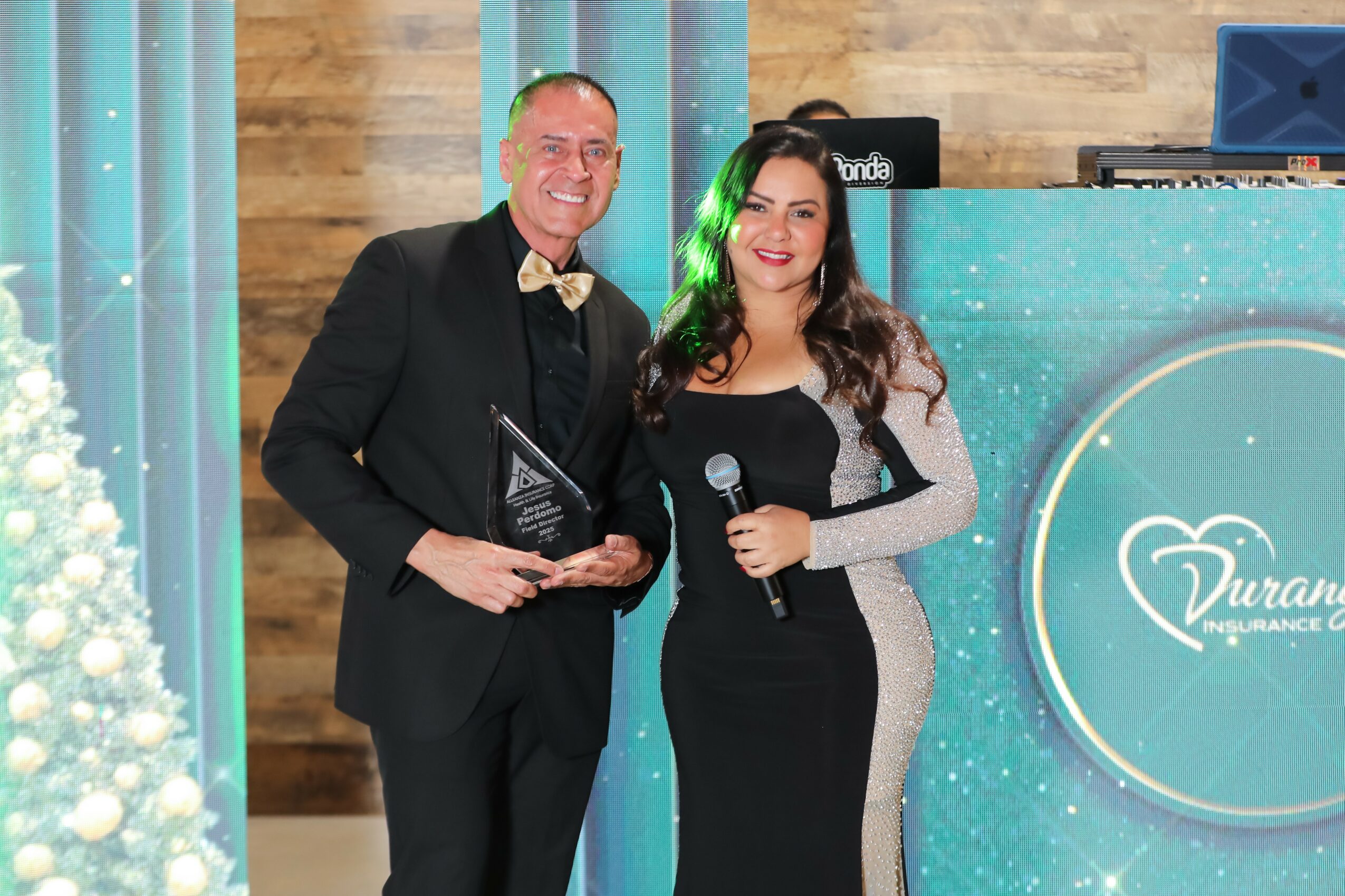 Man and woman standing together, smiling, holding an award and microphone, in a decorated event space with a logo background.