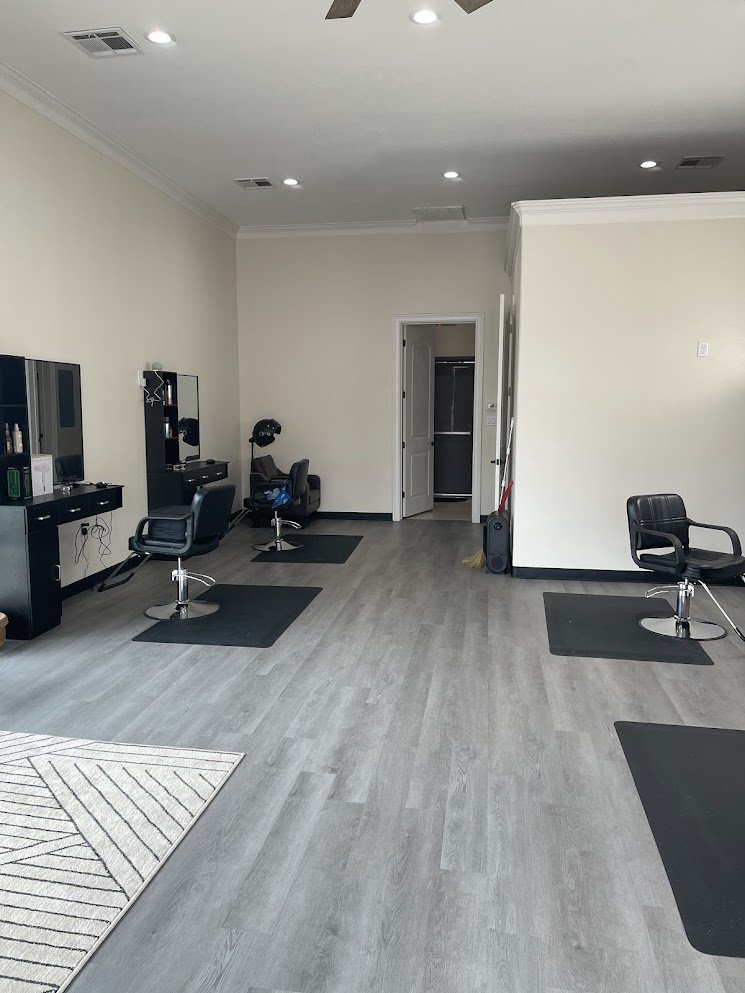 Empty hair salon with chairs, mirrors, and styling stations, light-colored walls, and wood-like flooring.