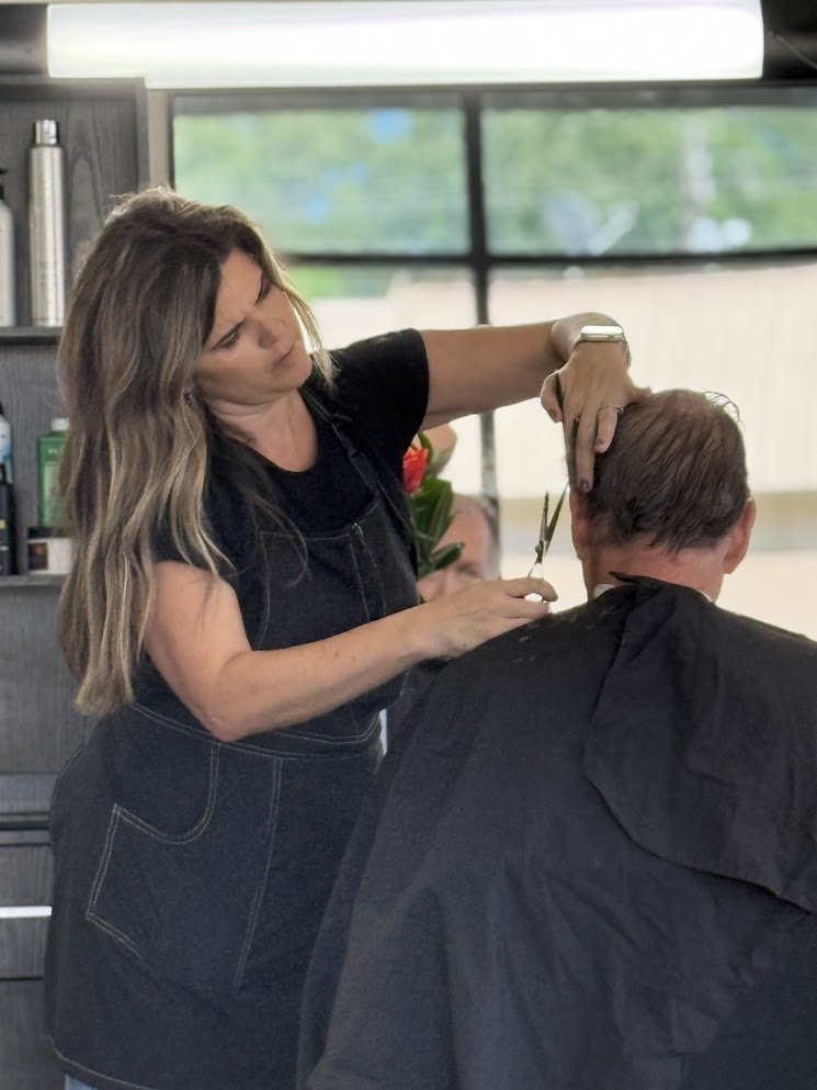 Hairdresser cutting a person's hair in a salon with large windows in the background.