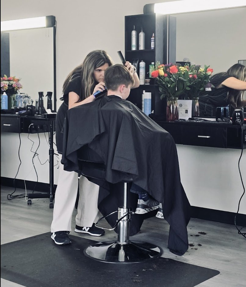 A person getting a haircut in a salon with a stylist working on their hair, mirrors, and salon supplies visible.