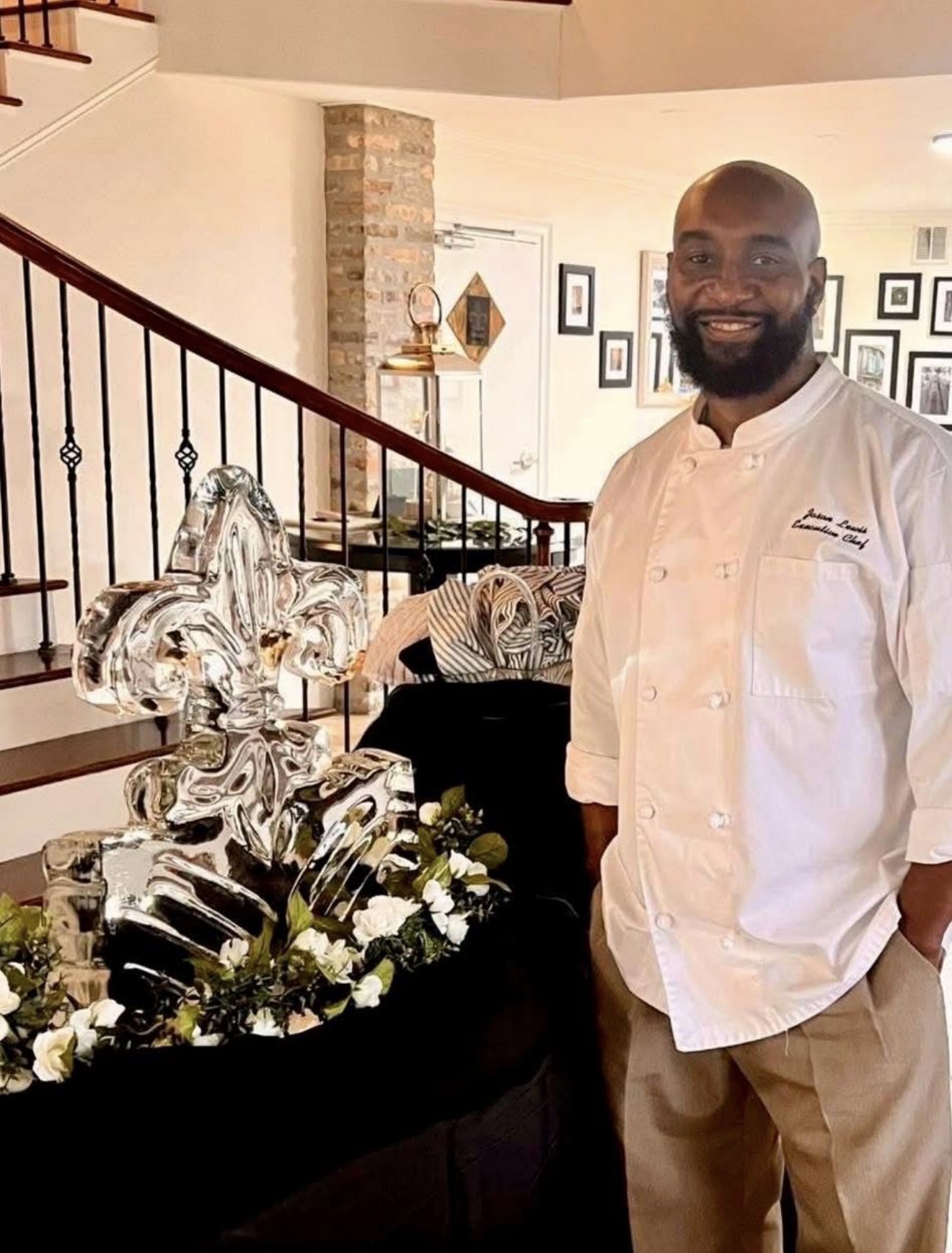 Man with beard in white chef's coat standing indoors near a table with decorative items and flowers, staircase in background.
