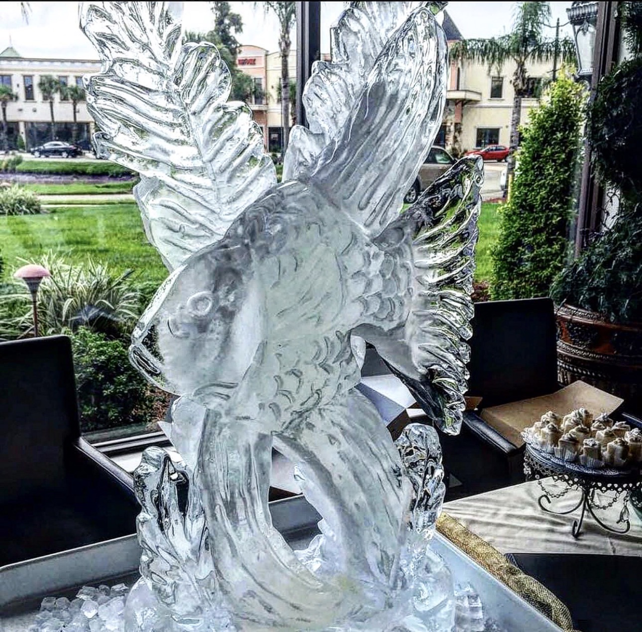 Ice sculpture of a fish with detailed fins and scales, displayed indoors near a window with a garden view.