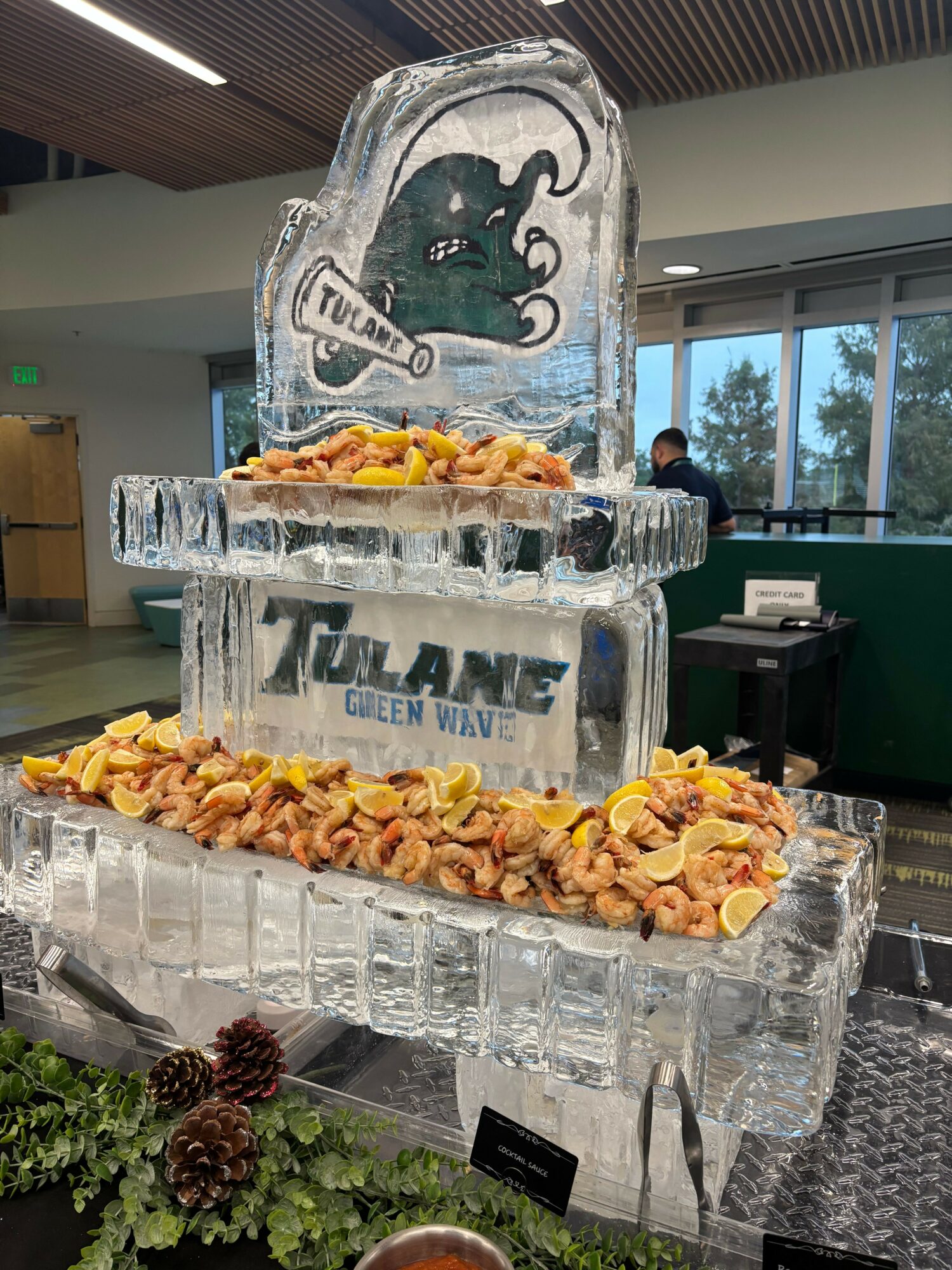 Ice sculpture with a logo, surrounded by flower petals, on a table with glasses and plants, indoors with windows.
