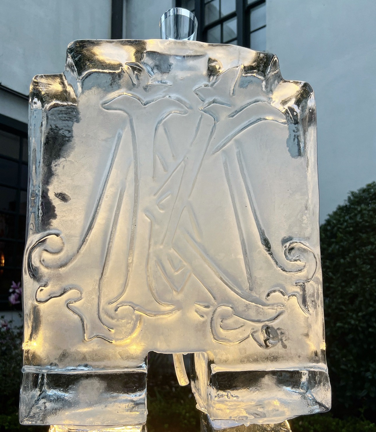 Ice sculpture with carved initials and decorative elements, outdoors with a building and trees in the background.
