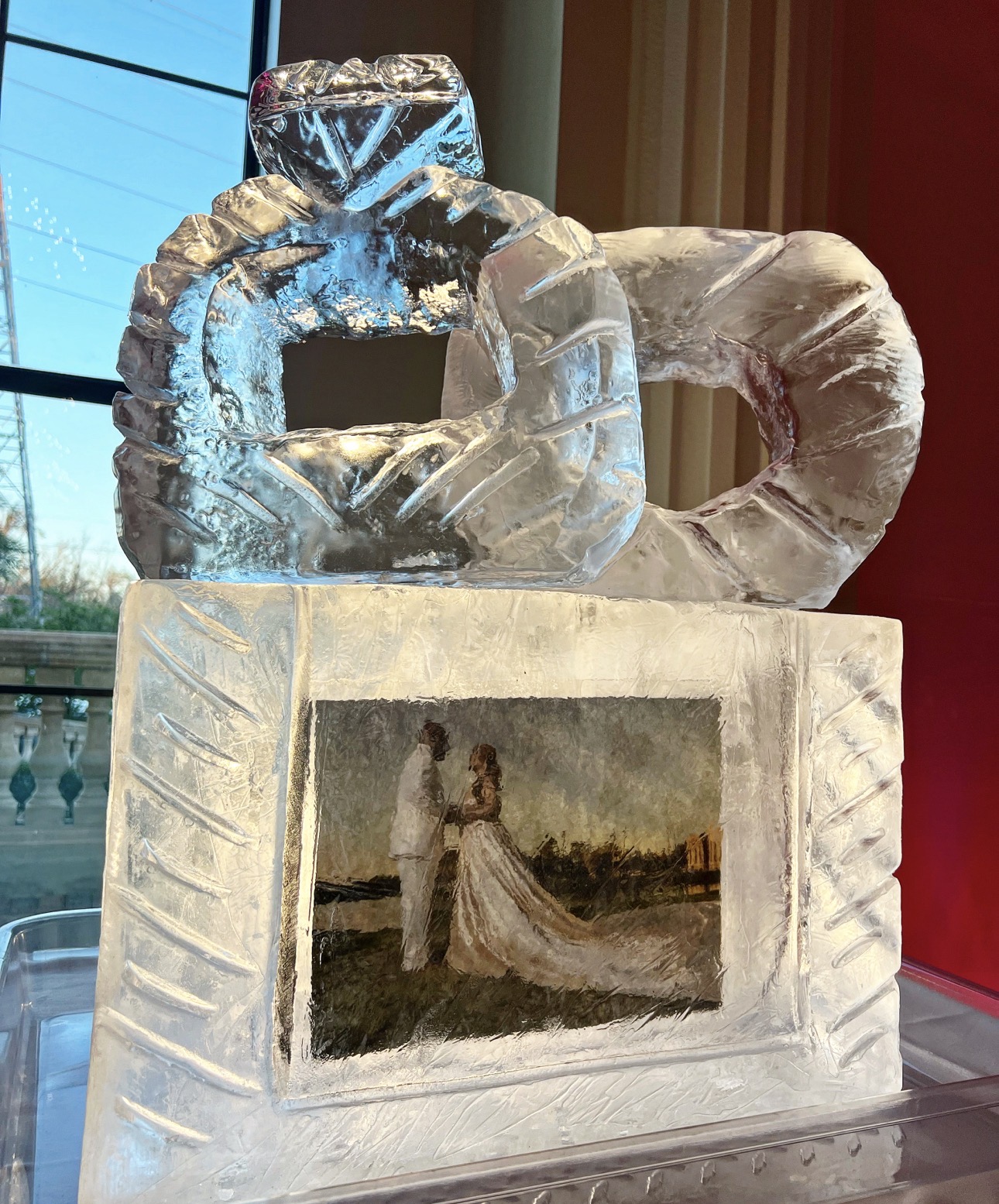 Ice sculpture of a camera with a painting of a woman in a dress inside, placed on a pedestal.