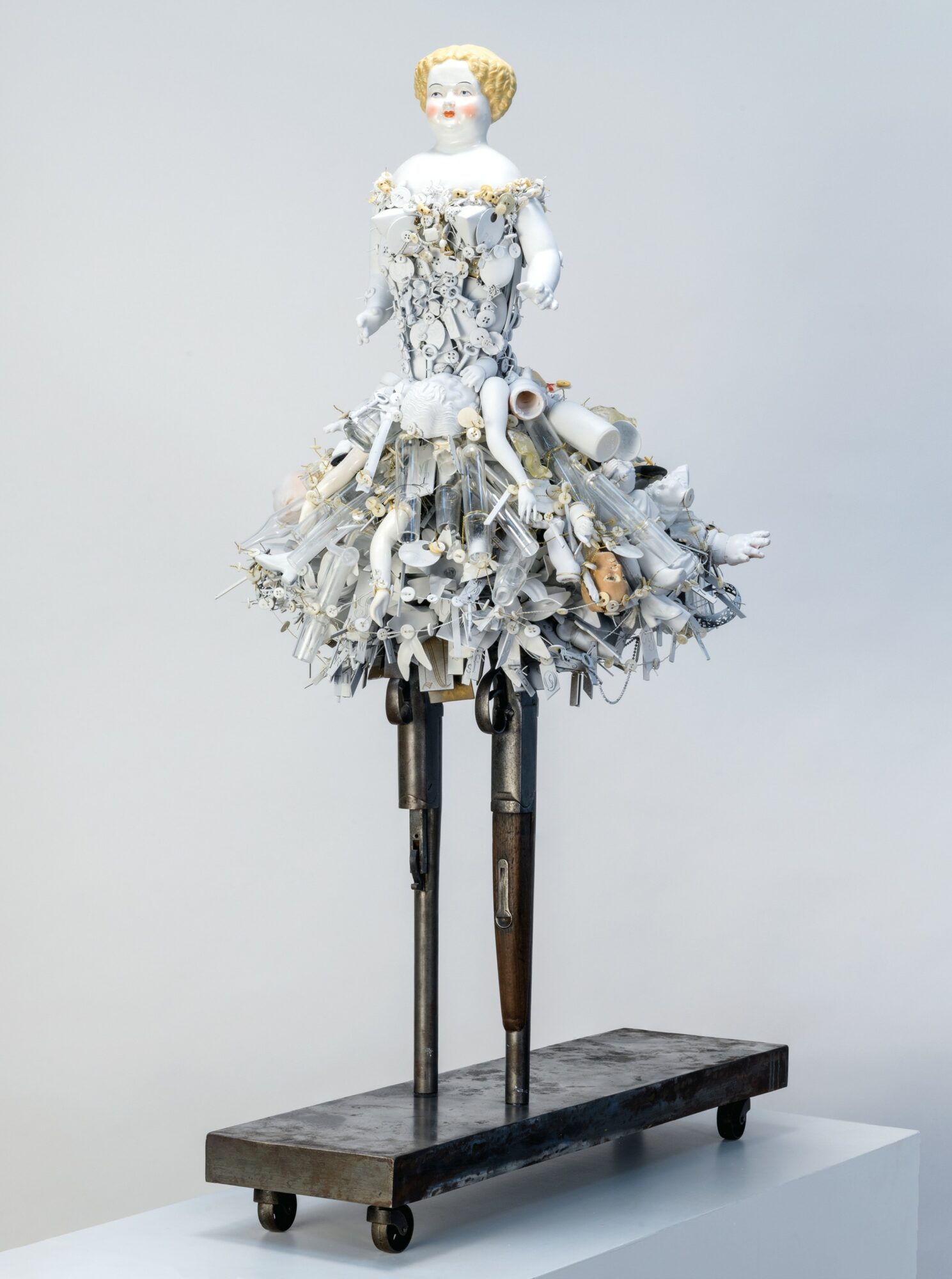 Sculpture of a doll with a dress made of various white and gray materials, mounted on tall legs on a wooden platform.