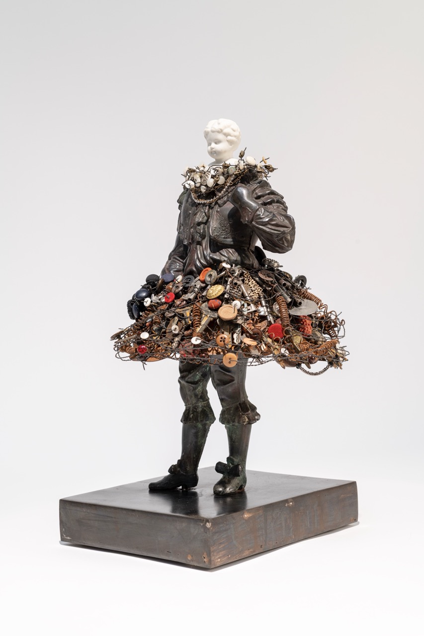 Sculpture of a figure with a textured dress made of various objects, standing on a rectangular base, against a plain background.