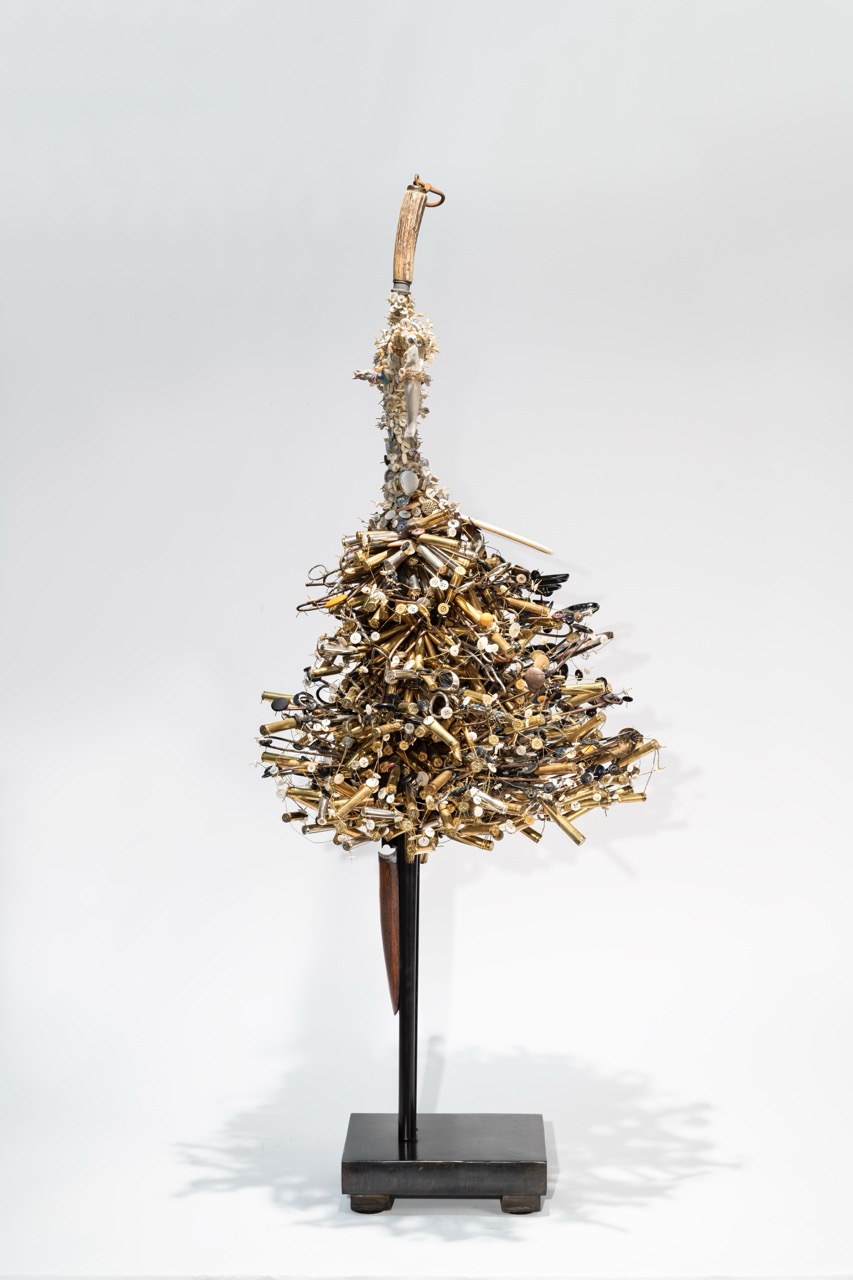 Sculpture resembling a tree made of metallic objects on a black stand, against a plain light background.