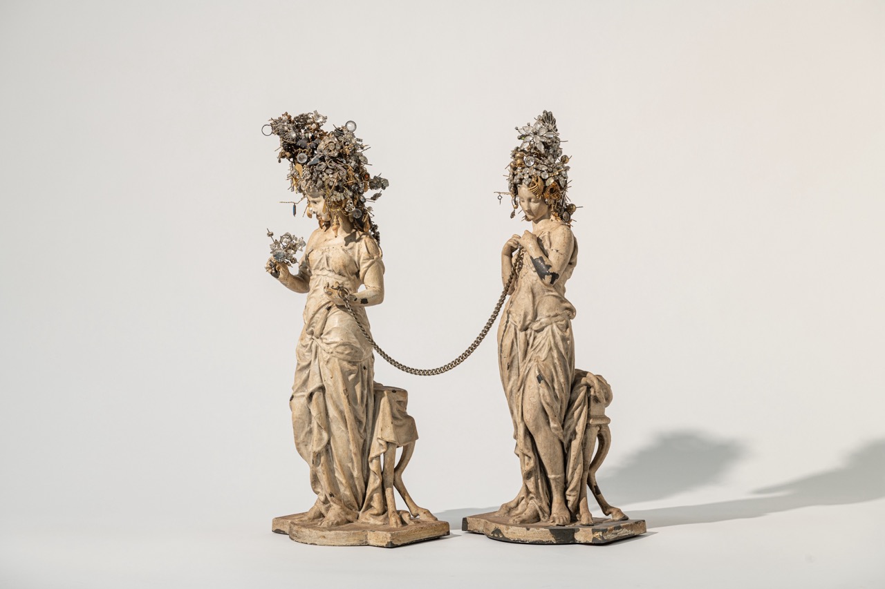 Two statues of women with floral crowns hold a rope between them, standing on a plain background.