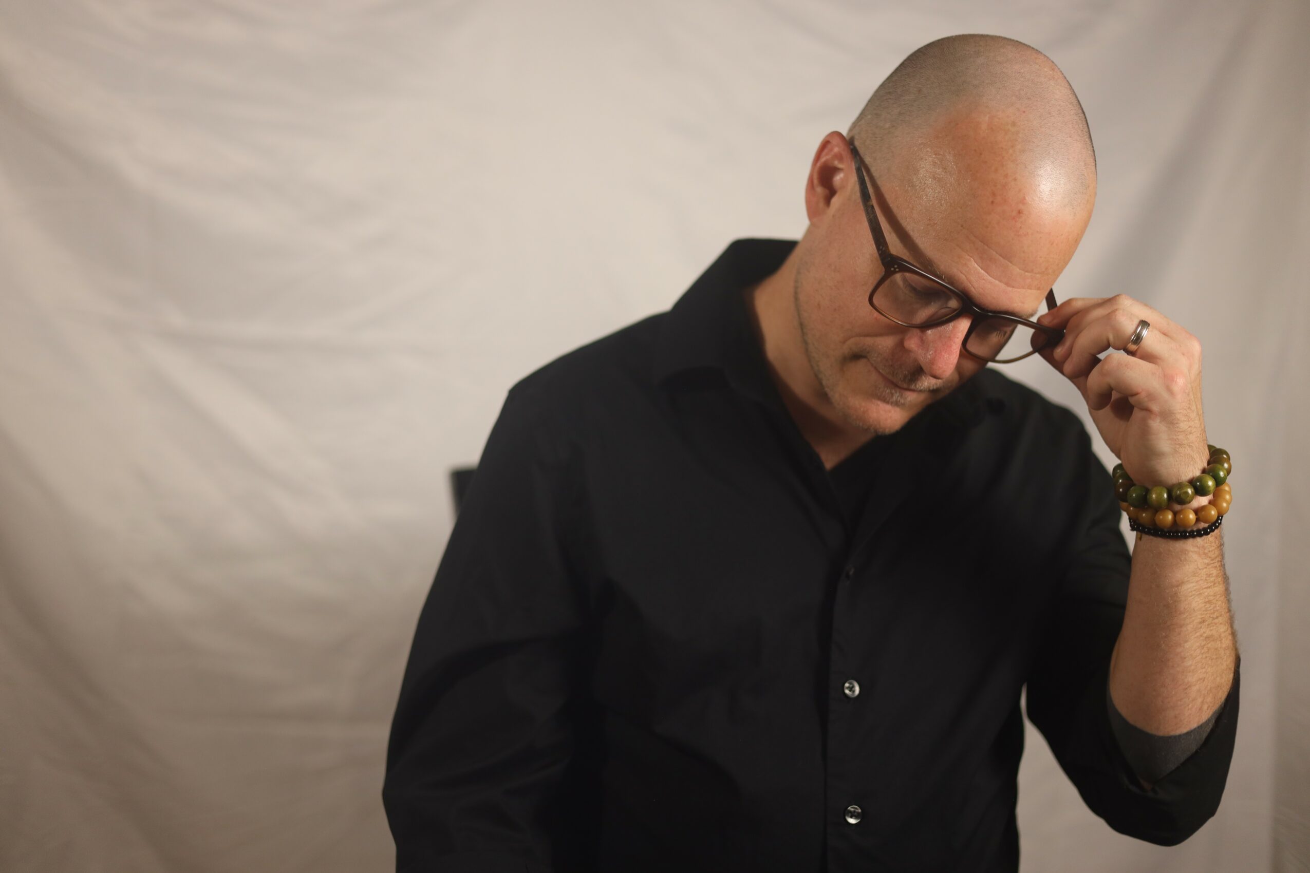 Bald man with glasses adjusting his glasses, wearing a black shirt, against a plain background.