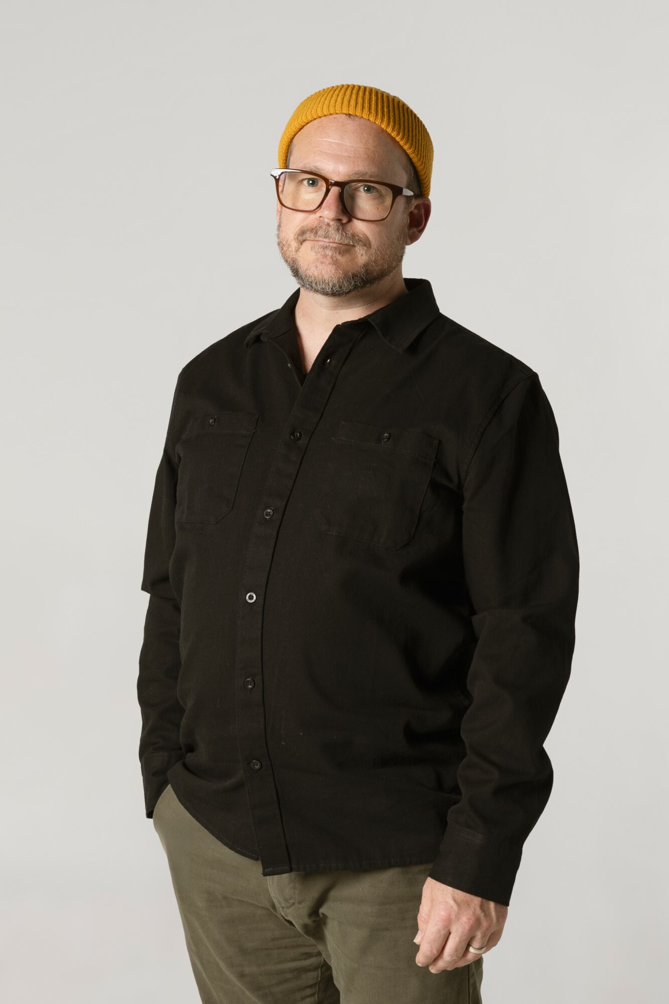 Man with glasses, beard, yellow beanie, black shirt, and beige pants stands against a plain background.