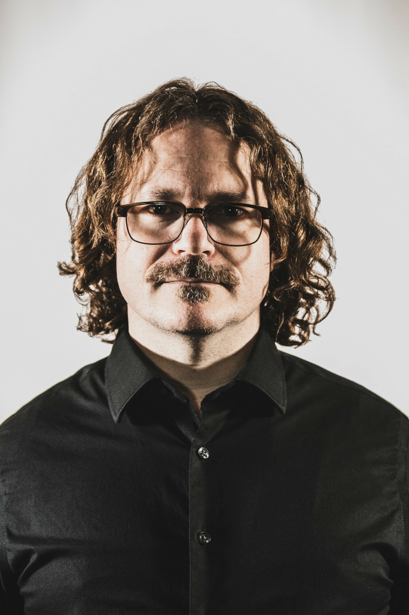 Man with curly hair, glasses, and a mustache wearing a black shirt, looking directly at the camera.