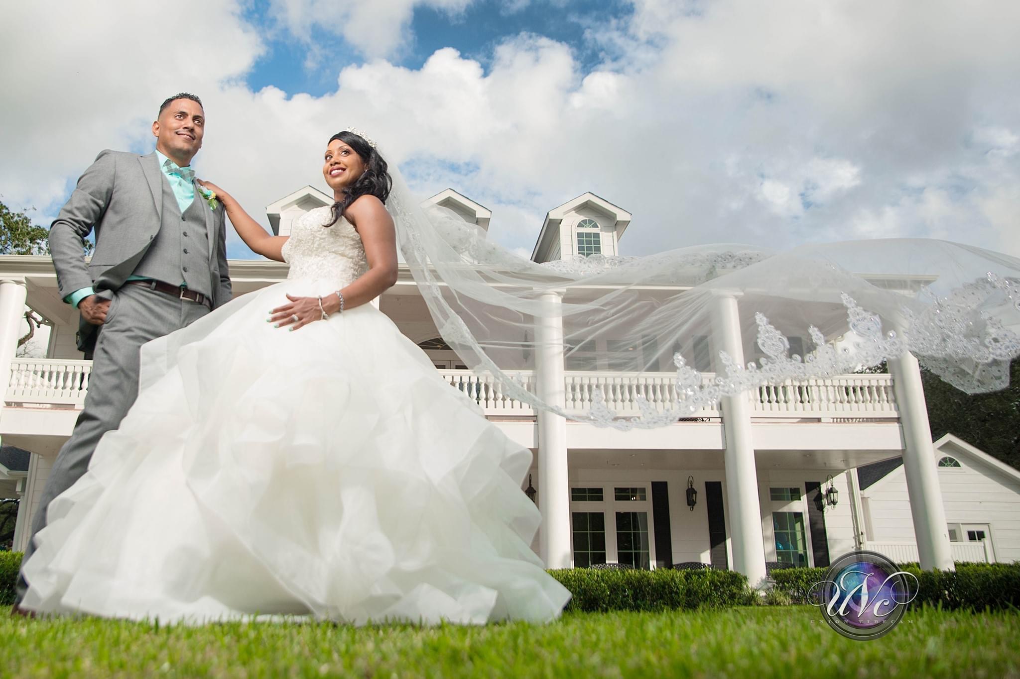 Bride and groom standing outside a large white house, bride in a wedding dress, groom in a suit, cloudy sky background.