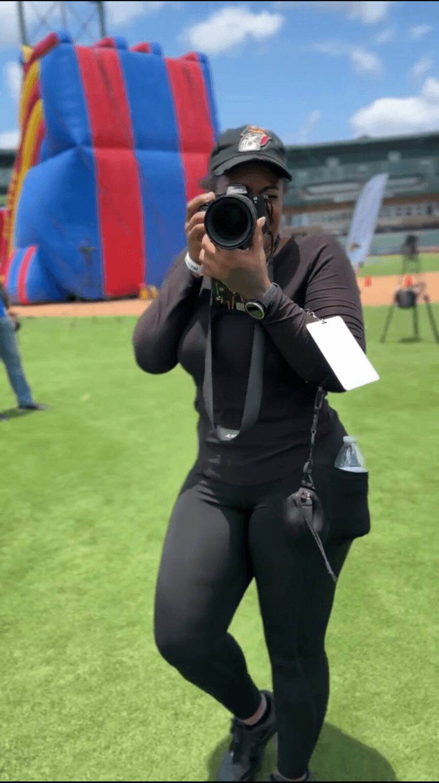 Person taking a photo with a camera outdoors, large inflatable slide in background, grassy field, cloudy sky.