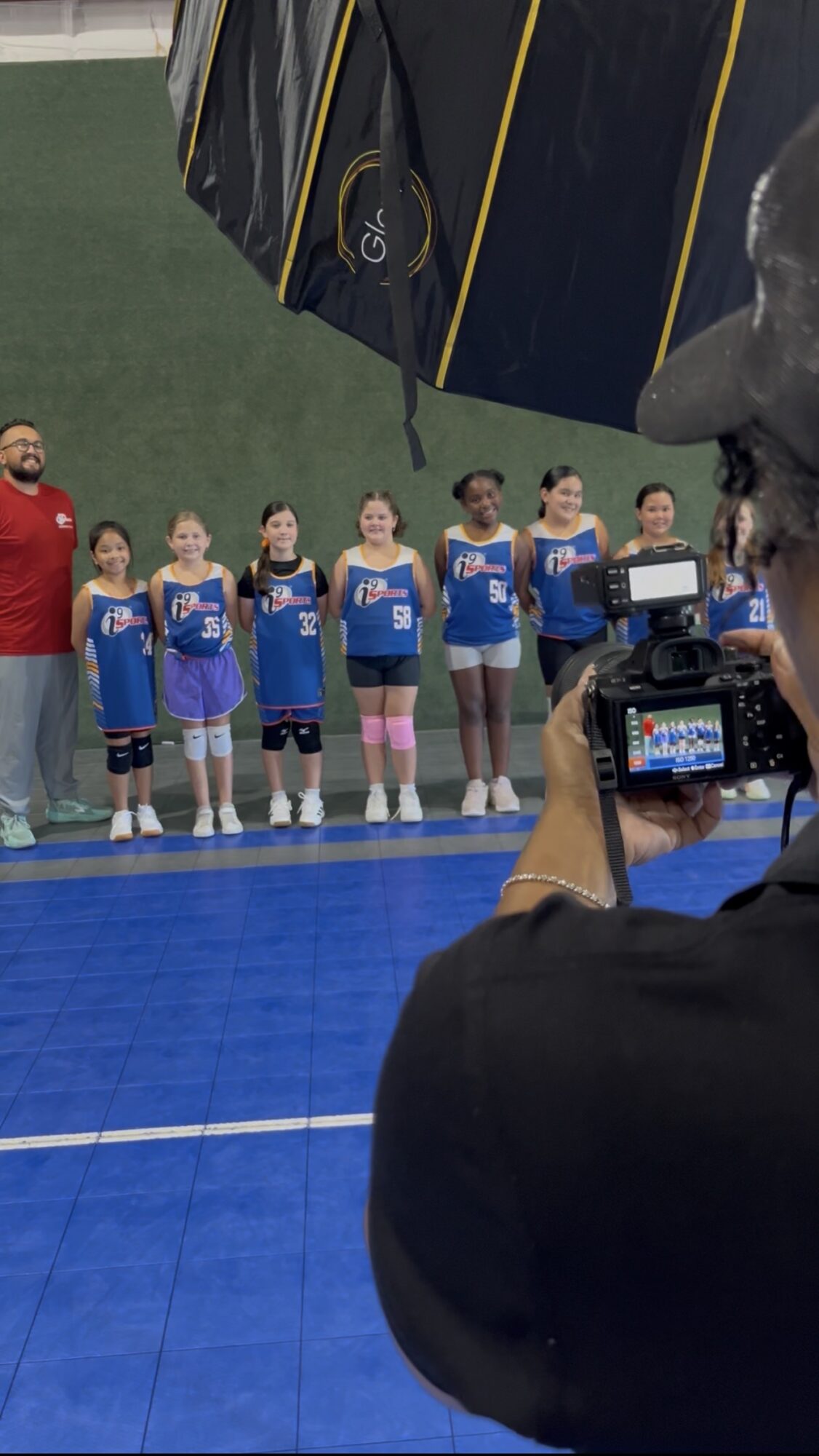 Group of young athletes standing on a blue indoor court, with a person taking a photo, and a large umbrella overhead.