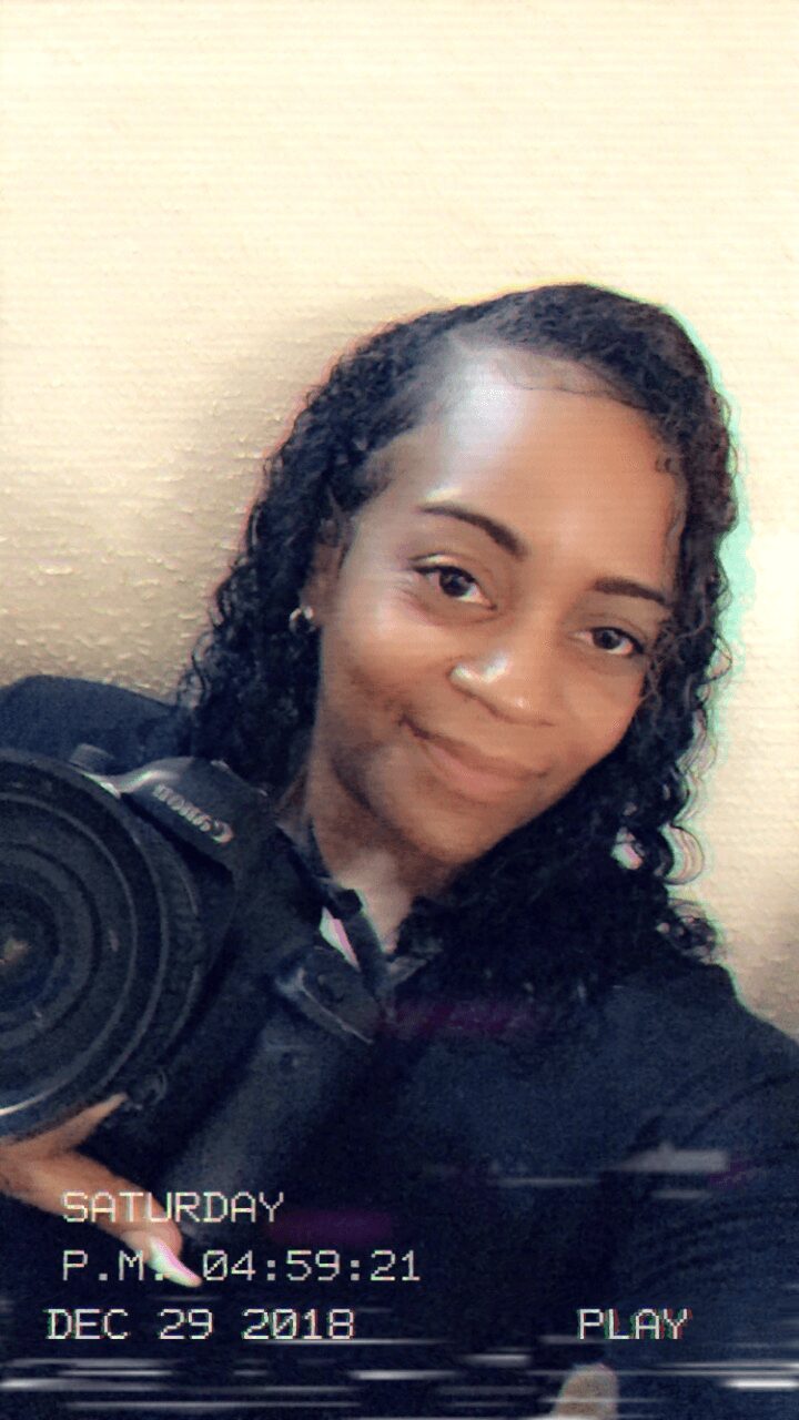 Young woman with curly hair smiling, holding a camera, against a plain background.