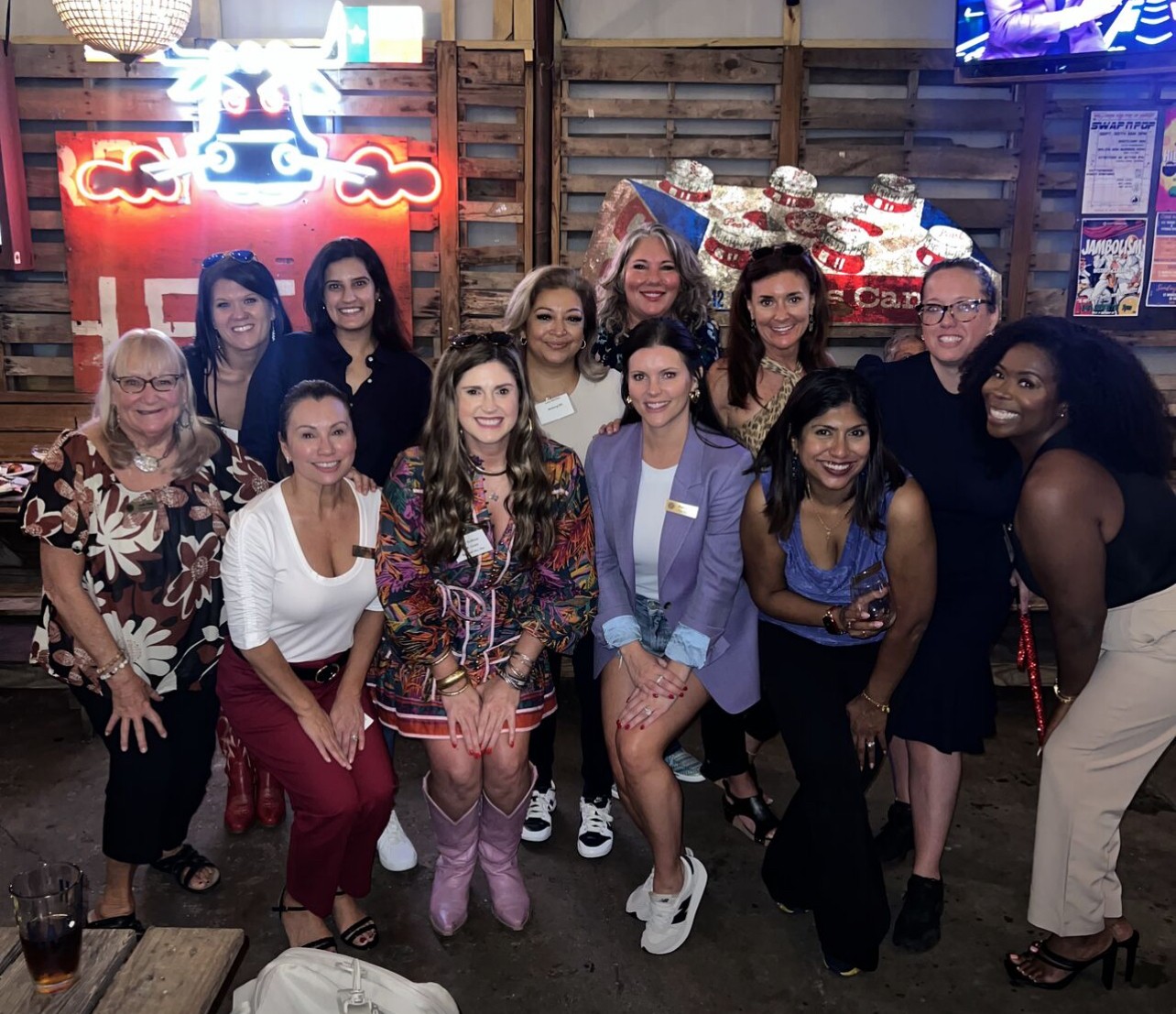 Group of twelve women and one man posing indoors with wooden walls and neon signs, smiling at the camera.