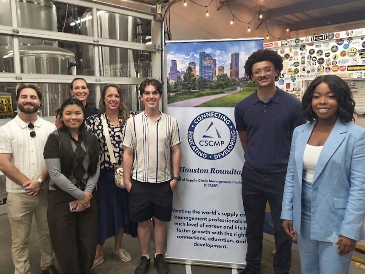 Group of seven diverse people standing indoors, smiling, with a banner and cityscape background, in a casual setting.