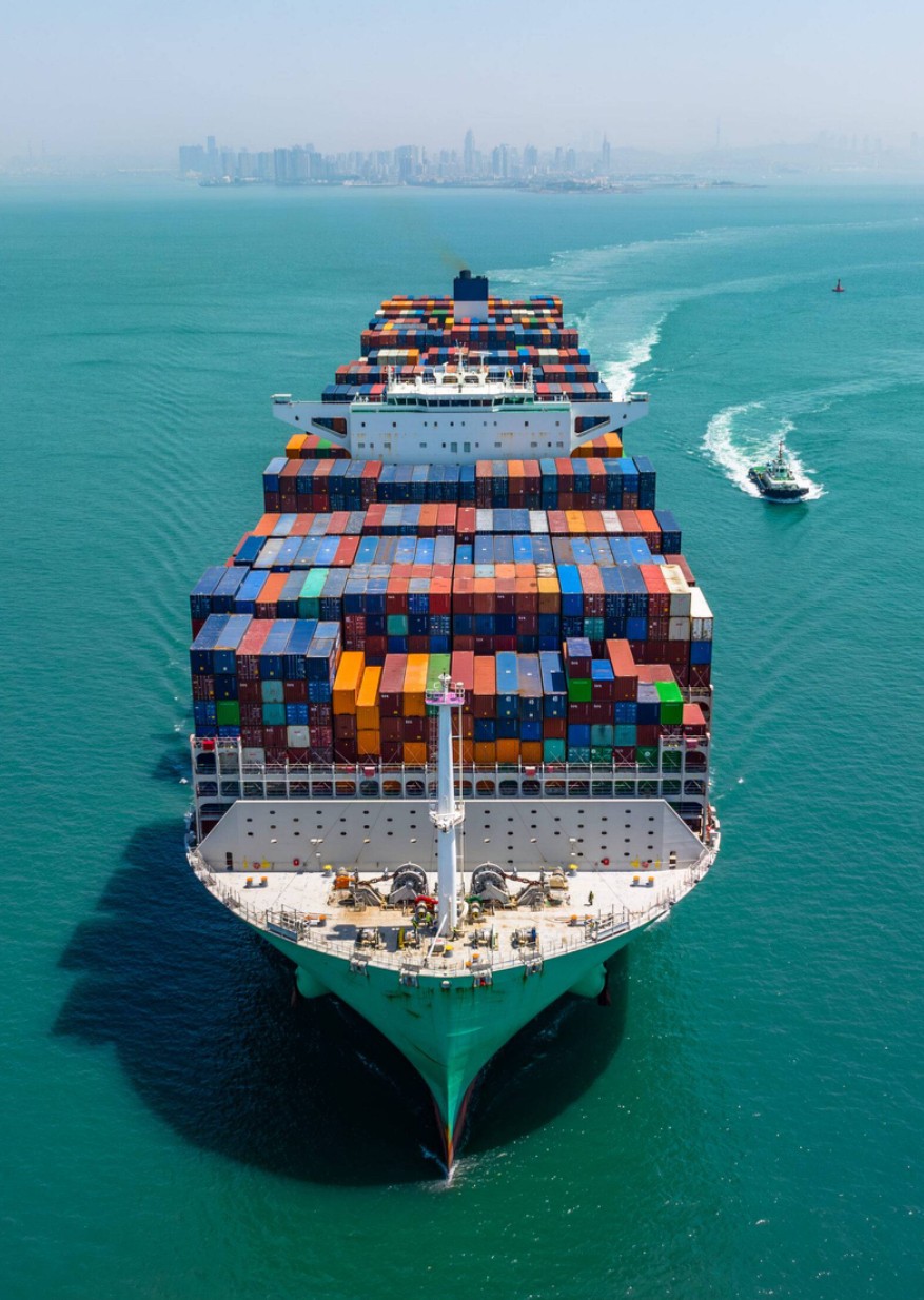 Large cargo ship with colorful containers sailing in open water, with a distant city skyline in the background.