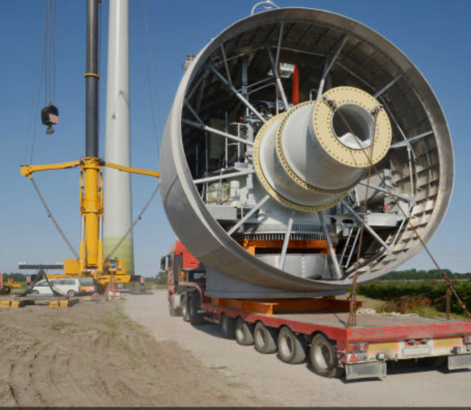 Large industrial equipment on a flatbed truck at a construction site with a crane nearby, under a clear blue sky.