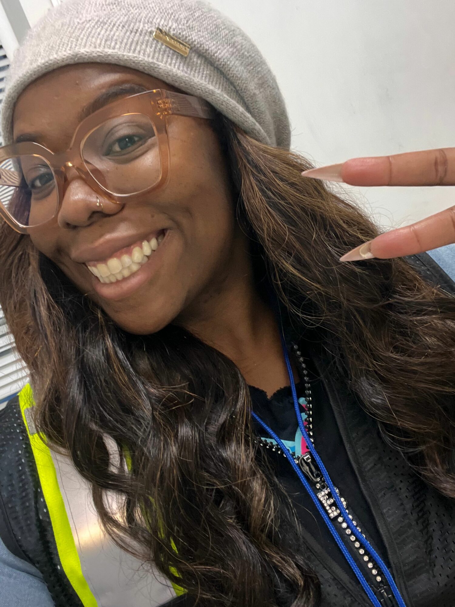 Person smiling, wearing glasses and a gray beanie, making a peace sign with fingers, with long dark hair and a black jacket.