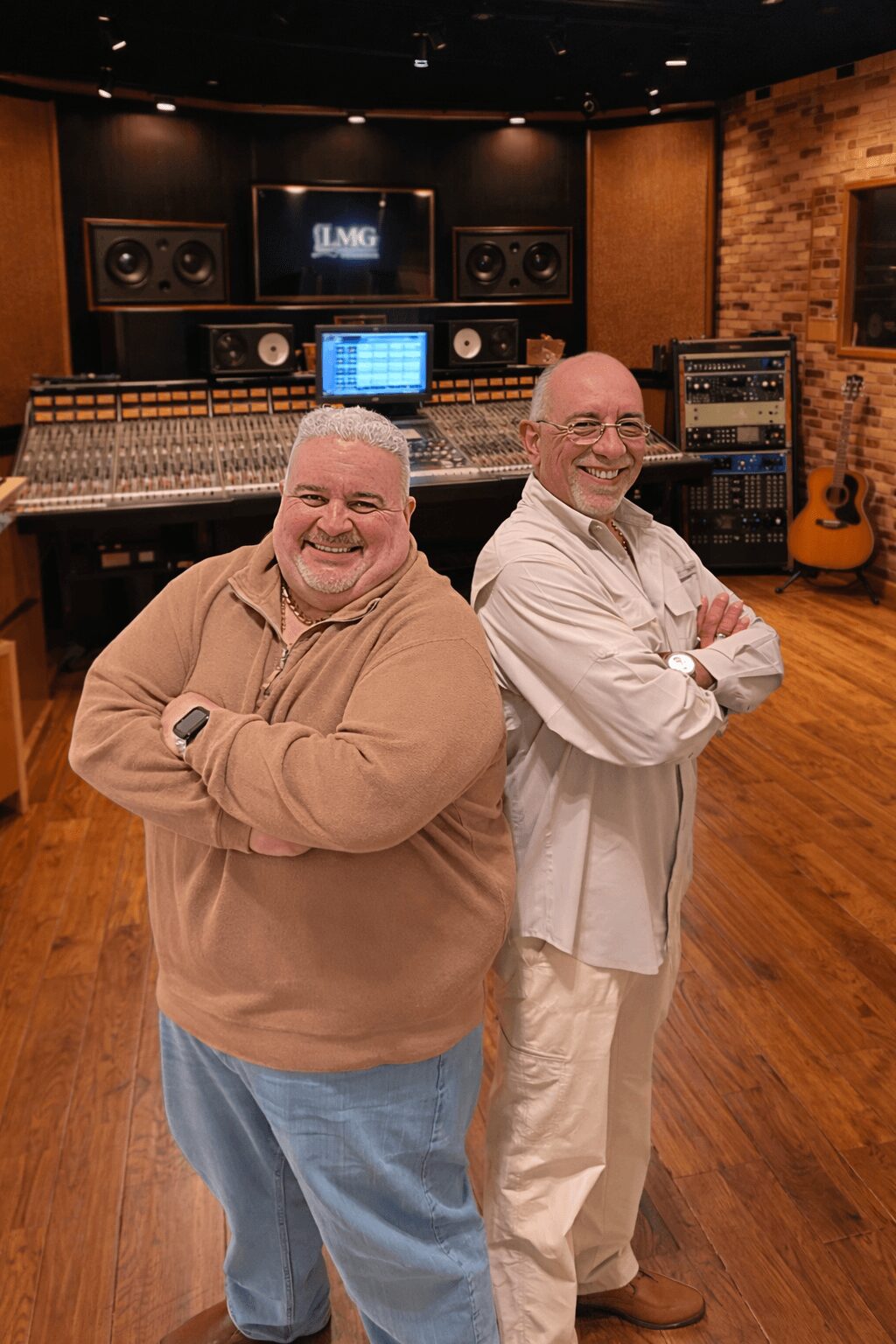 Two smiling men stand with arms crossed in a recording studio with audio equipment and guitars in the background.
