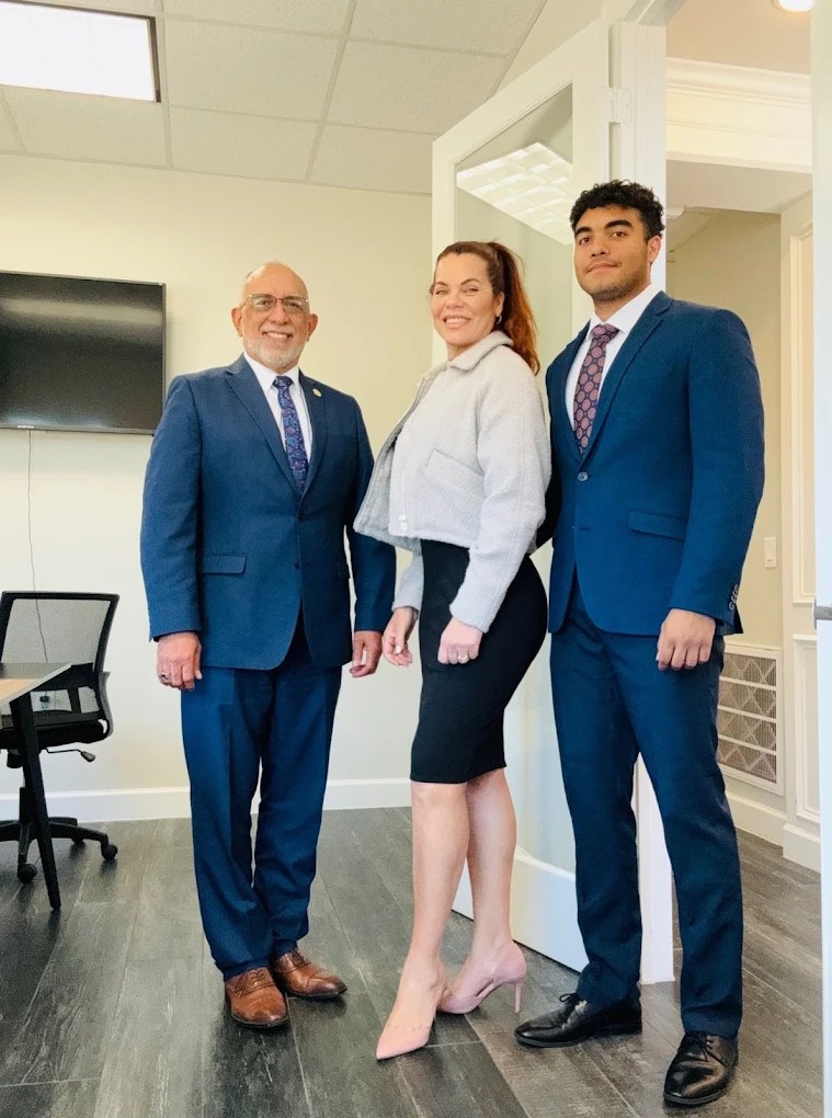 Three professionally dressed people standing in an office, smiling, with a TV and office chair in the background.