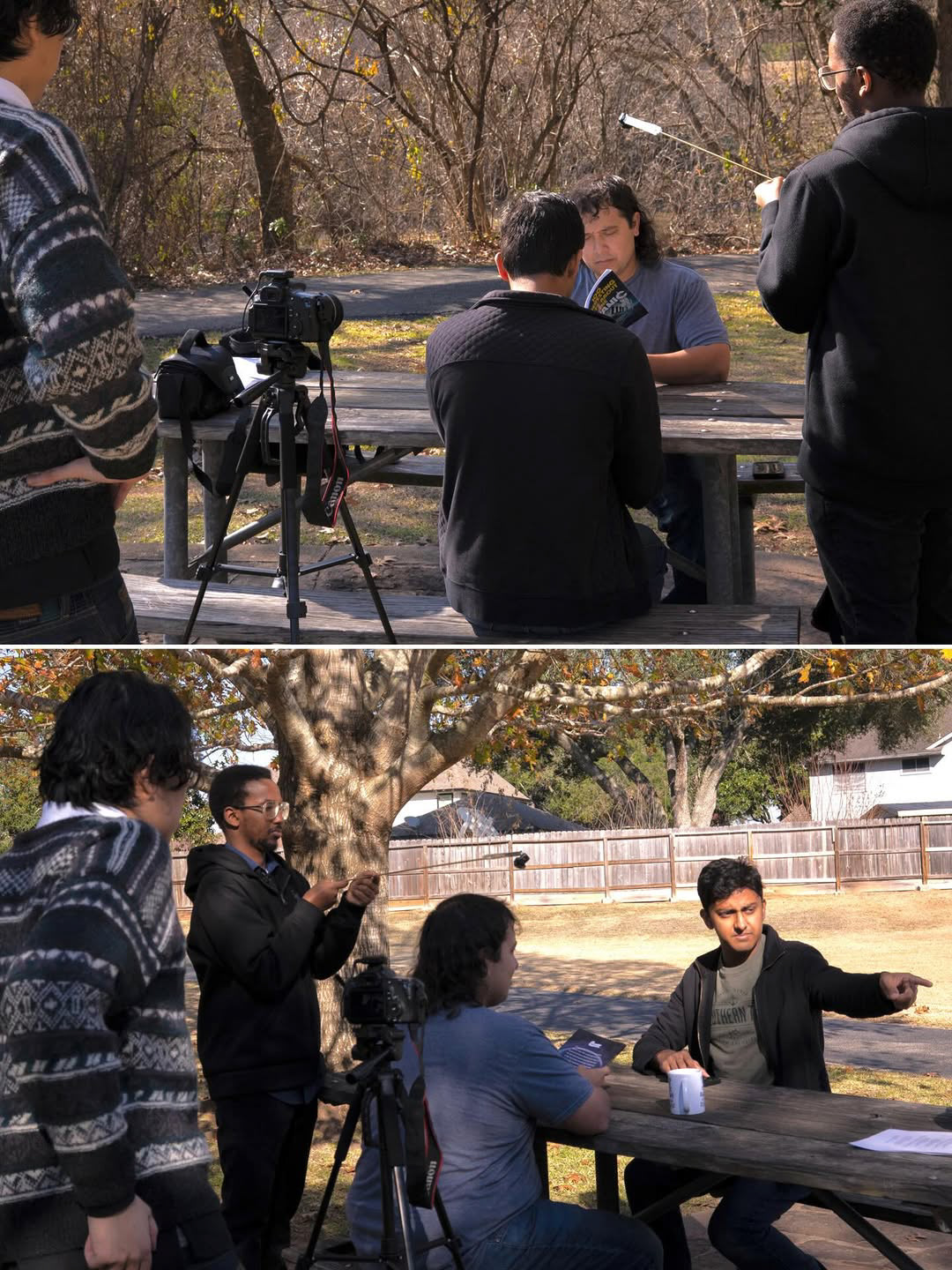 Group of people outdoors, some sitting at a table, others standing, with cameras and equipment, trees and a fence in background.