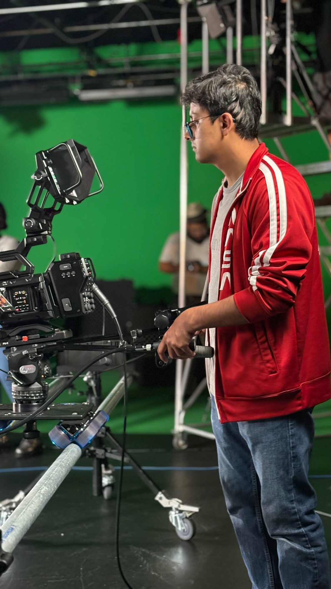 Young man in red jacket operating camera on studio set with green screen background.