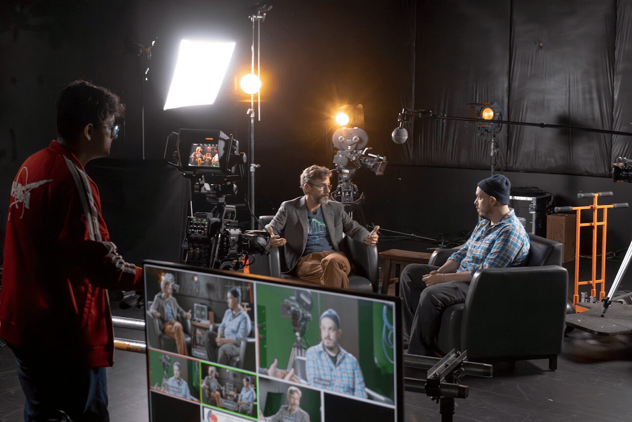 Two men sit in chairs on a film set with cameras and lighting, while a crew member operates a camera. Studio background visible.