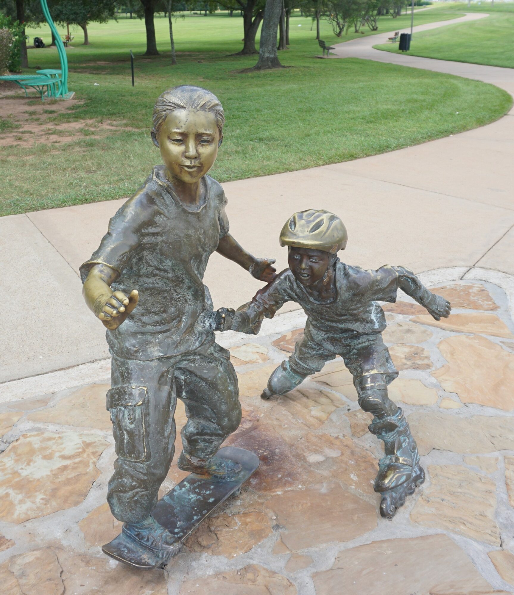 Bronze sculpture of two children, one girl and one boy, holding hands and skating on a paved area in a park.