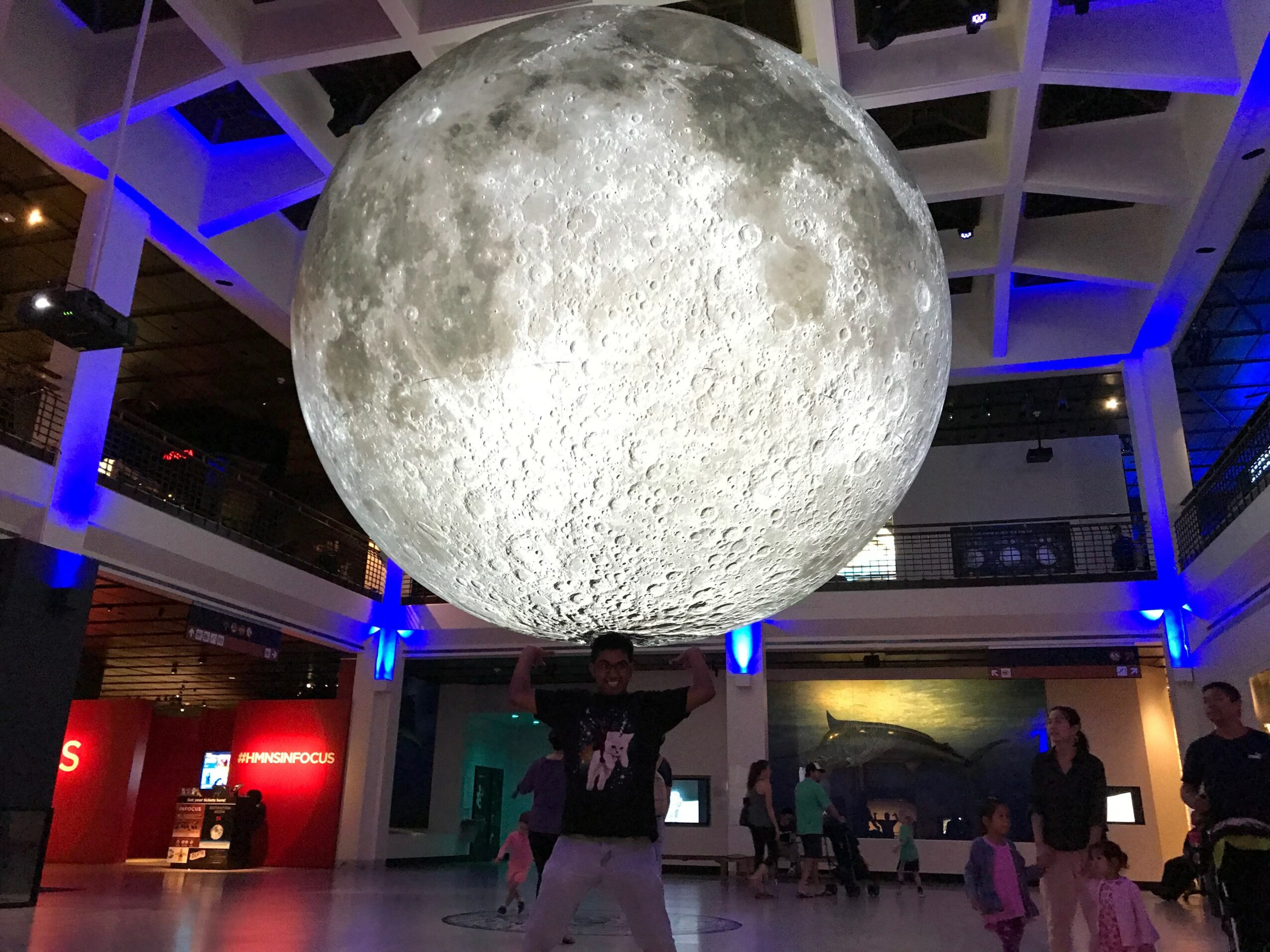 Large moon model hanging from ceiling in a modern indoor space with people and blue lighting.