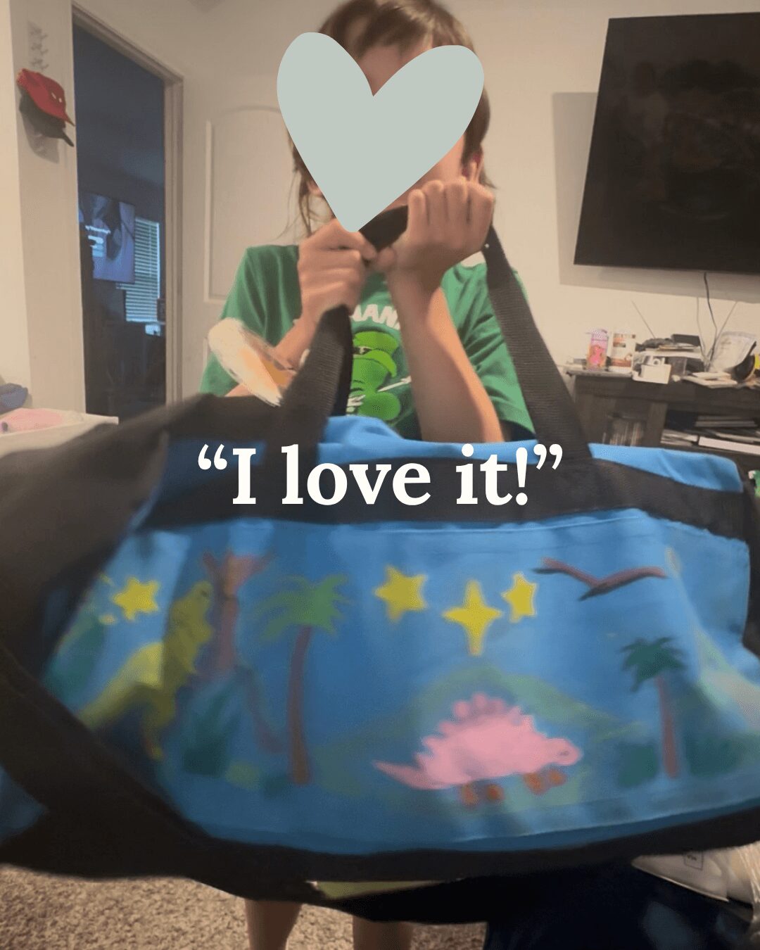Person holding a camera with a heart overlay, sitting behind a colorful bag with dinosaurs and stars, in a room with a TV and door.