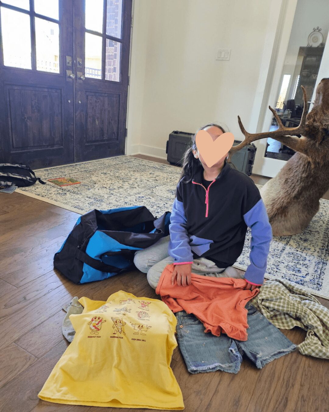 Child sitting on the floor with clothes and a backpack, in a room with wooden floor and large window, face obscured.