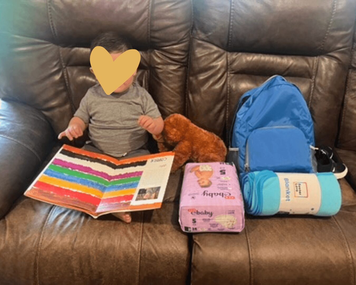 Child sitting on a brown leather couch reading a colorful book, with a small brown dog, a blue backpack, and a pink pack of diapers nearby.