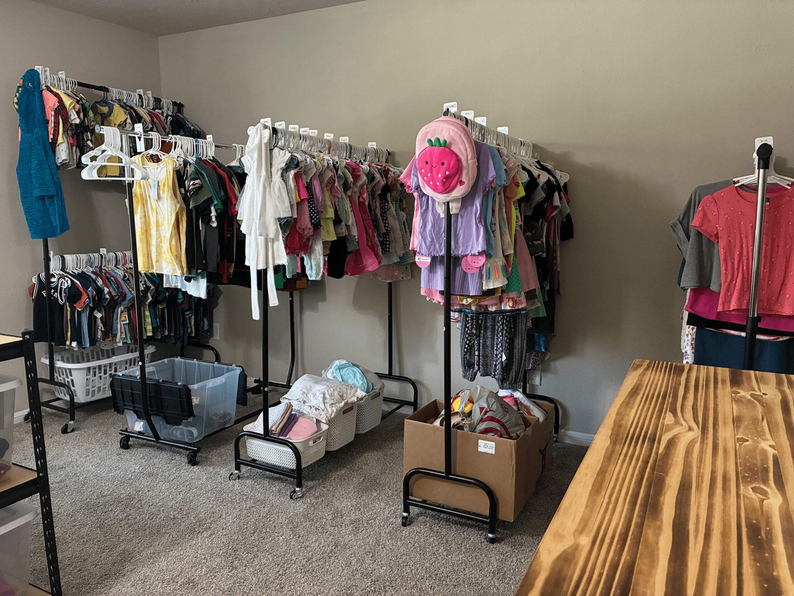 Clothing racks with children's clothes, a pink backpack, and boxes of clothes in a room.
