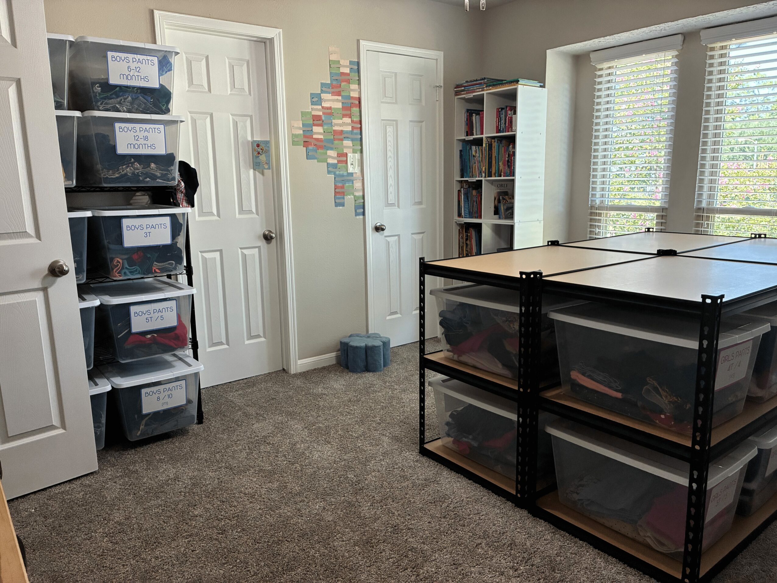 Room with shelves, storage bins, a table, and windows with blinds, carpeted floor, and a door in the background.