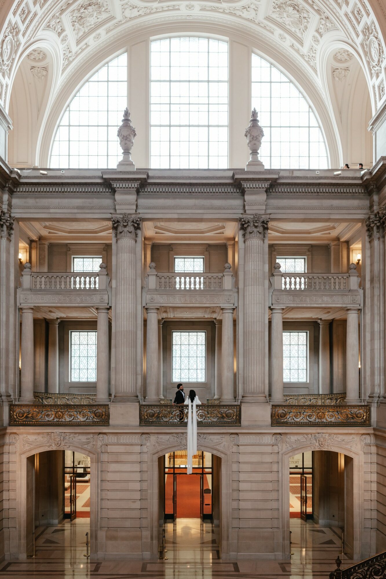 Interior of a grand building with tall windows, ornate architecture, and a person standing on a balcony overlooking the space.